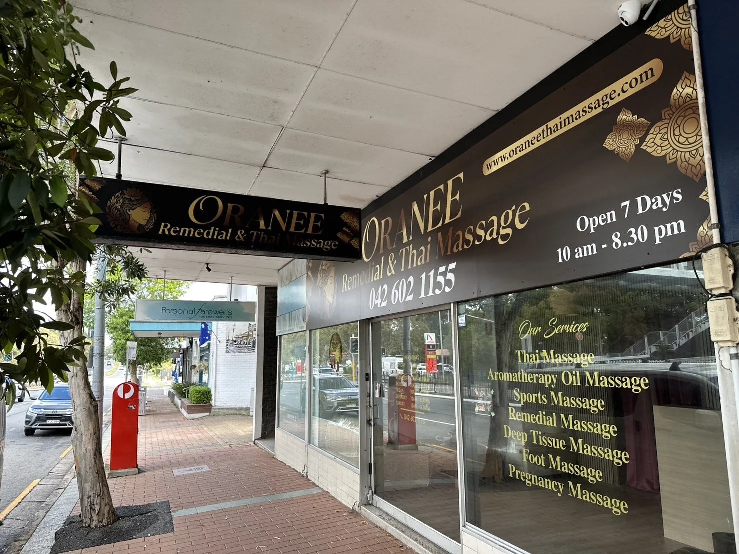 The storefront of ORANEE Remedial & Thai Massage, with large signs displaying services and open hours. The sidewalk in front has a tree, a fire hydrant, and parked cars in the background.