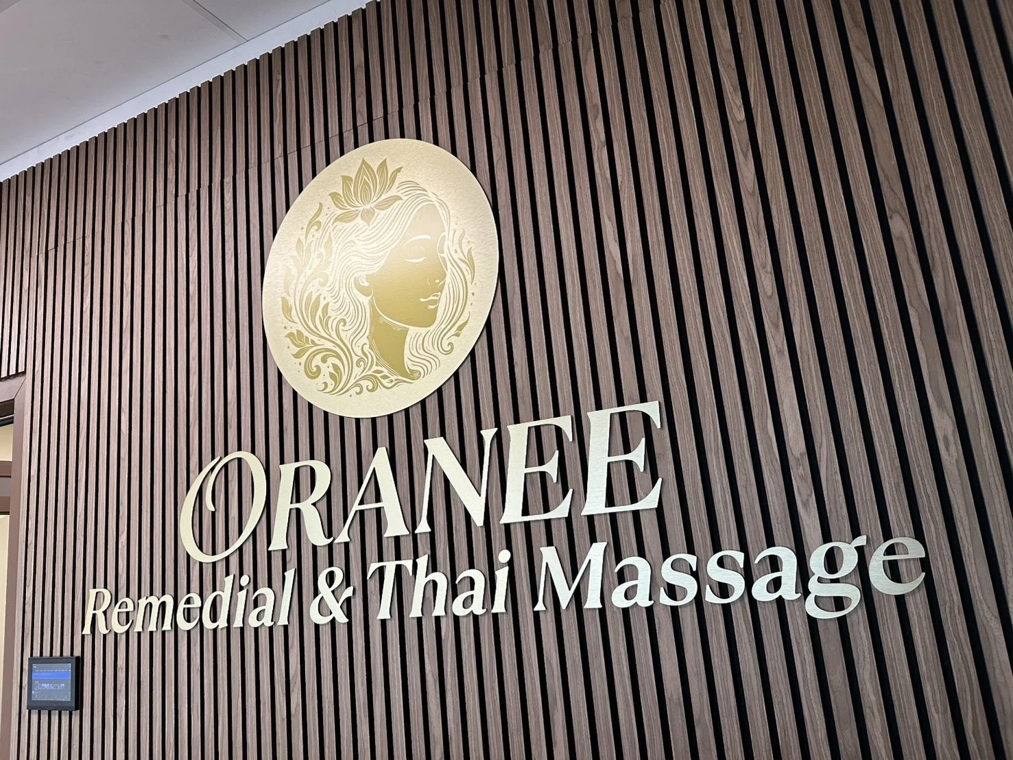 Wood-paneled wall with a circular logo of a woman's face surrounded by floral designs, above the sign 'ORANEE Remedial & Thai Massage'.