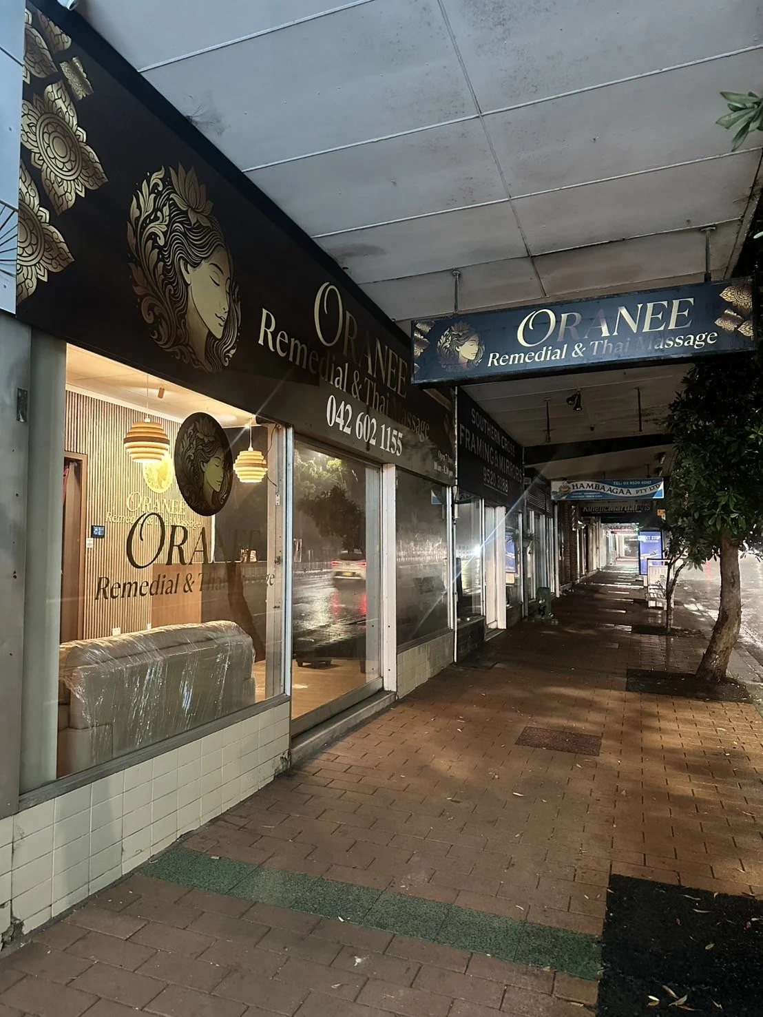 Exterior of a storefront for ORANEE Remedial & Thai Massage, with a black sign featuring a blonde woman’s face in gold outline, large windows, and a brick sidewalk with trees.