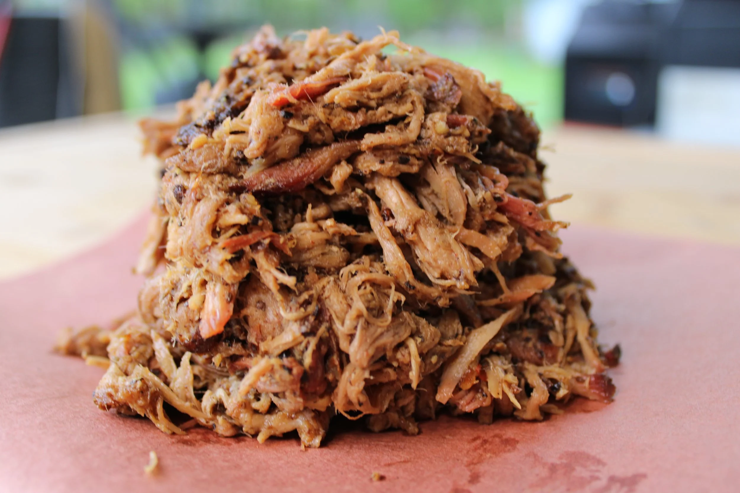 A close-up of a pile of shredded cooked meat on pink parchment paper, with a blurred outdoor background.