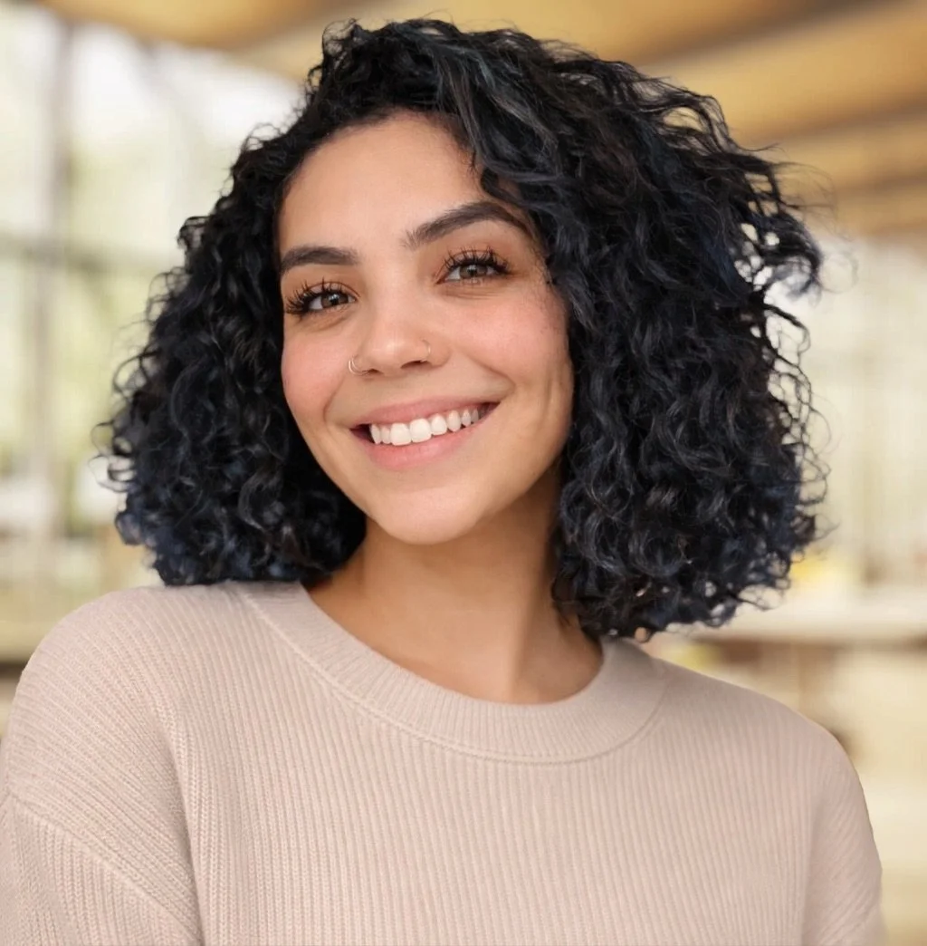 A young woman with curly black hair, smiling, wearing a beige top, in an indoor setting with blurred background.