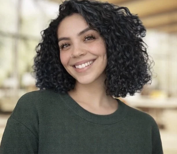 A young woman with curly black hair, smiling, wearing a dark green shirt indoors.