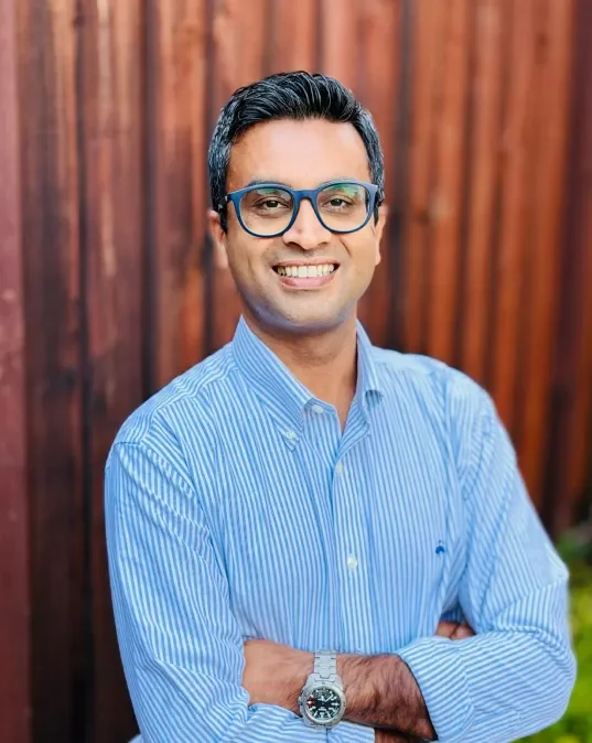 A smiling man in a blue striped button-up shirt, wearing glasses and a watch, stands with arms crossed in front of a wooden fence.
