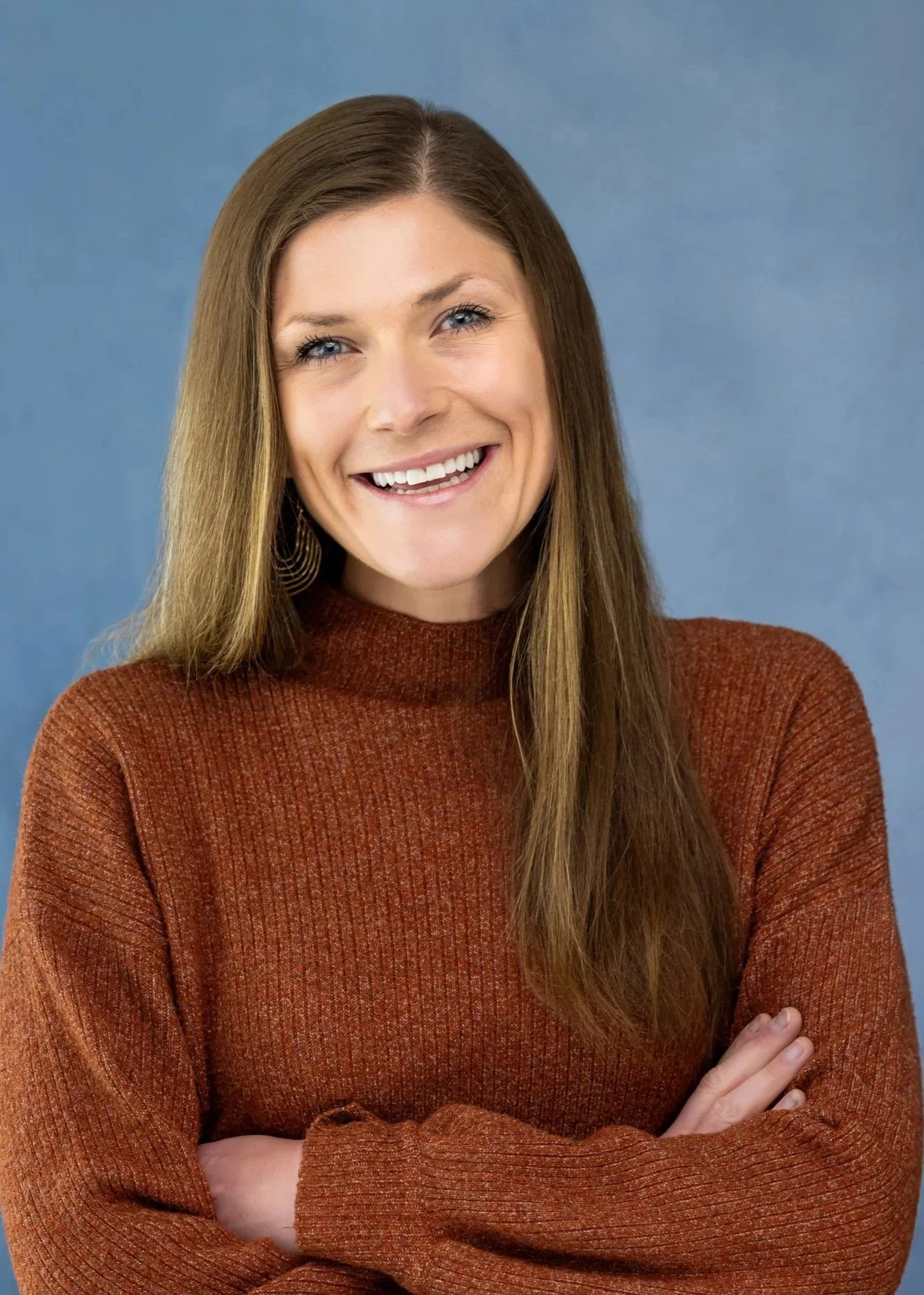 A smiling woman with long brown hair, wearing a rust-colored sweater, standing with her arms crossed against a blue background.