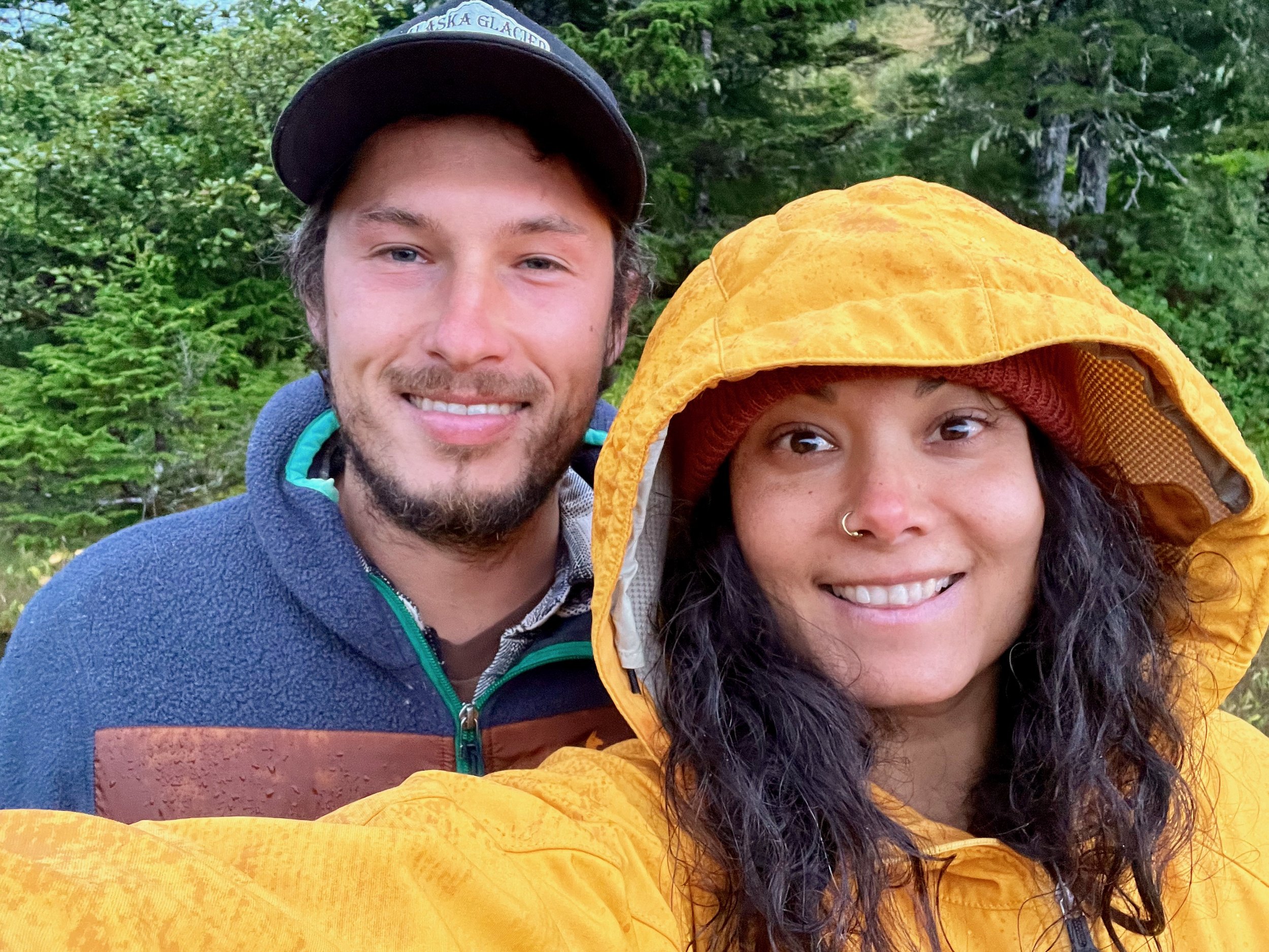 A young man and woman taking a selfie outdoors in a forest, dressed in jackets; the woman is wearing a yellow raincoat with the hood up and has a nose piercing. The man is wearing a dark cap and a fleece jacket.