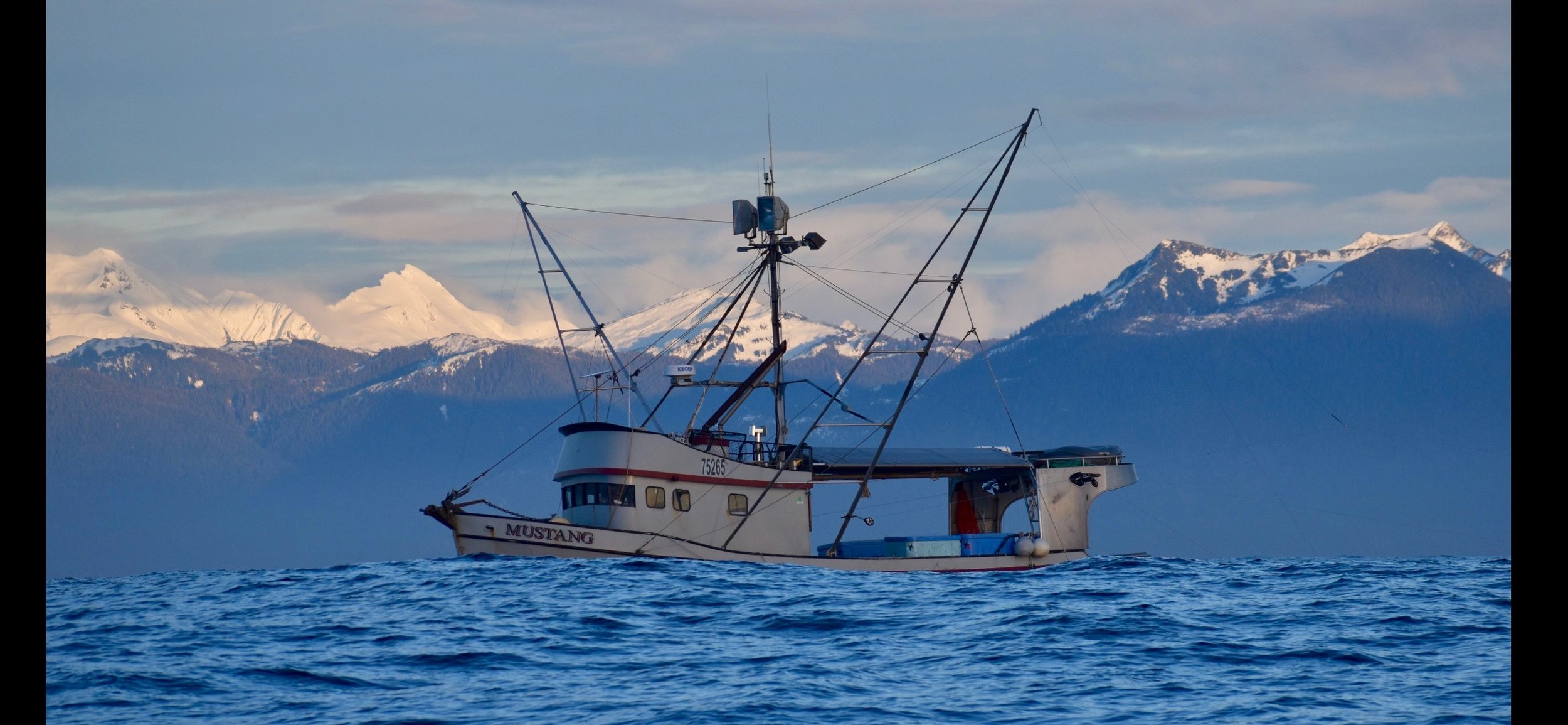 A fishing boat named Mustang on the water with snow-capped mountains in the background.