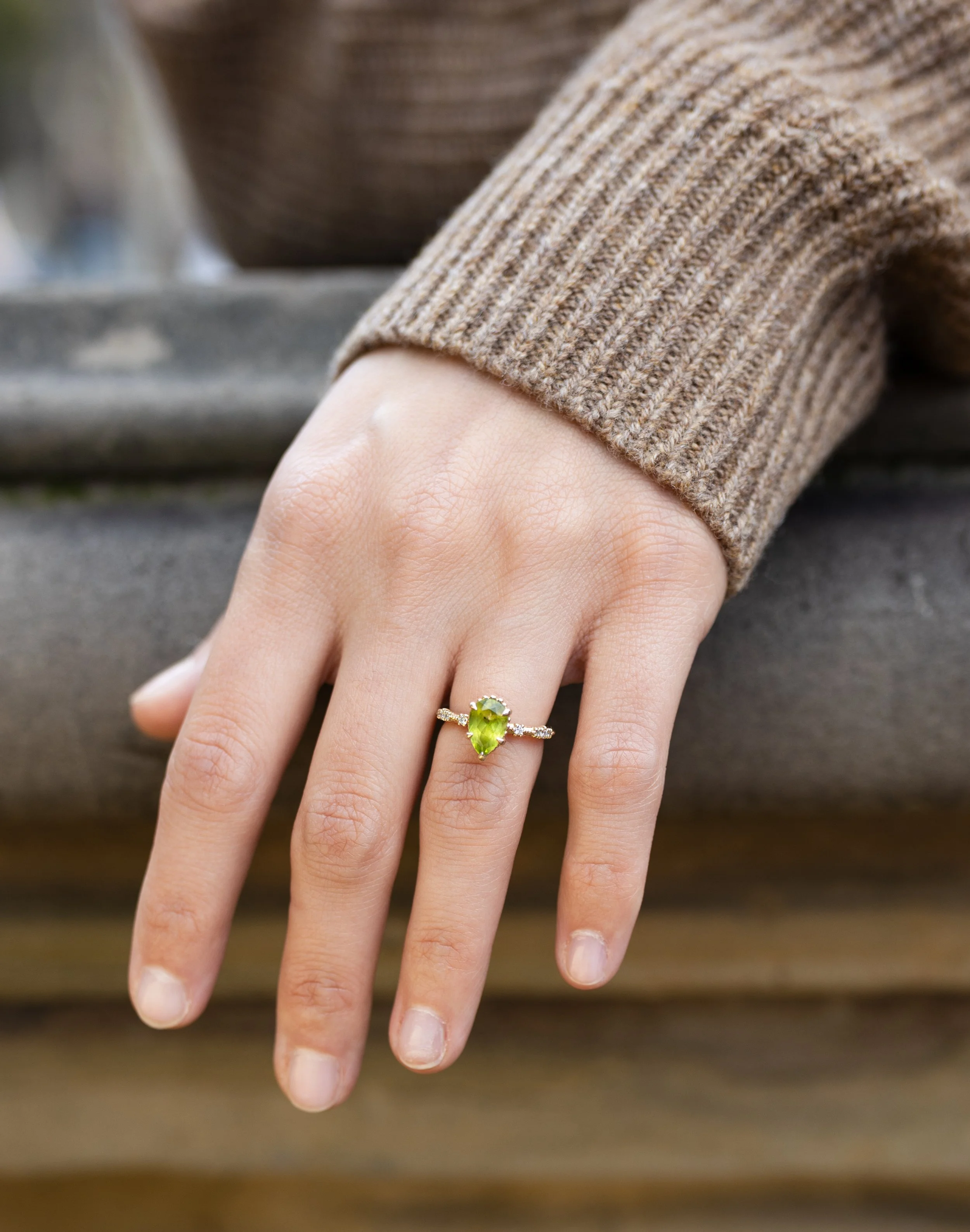 Close-up of a woman's hand wearing an engagement ring with a teardrop-shaped green gemstone and small diamonds at the band, resting on a wooden surface, with a brown knitted sweater sleeve visible.
