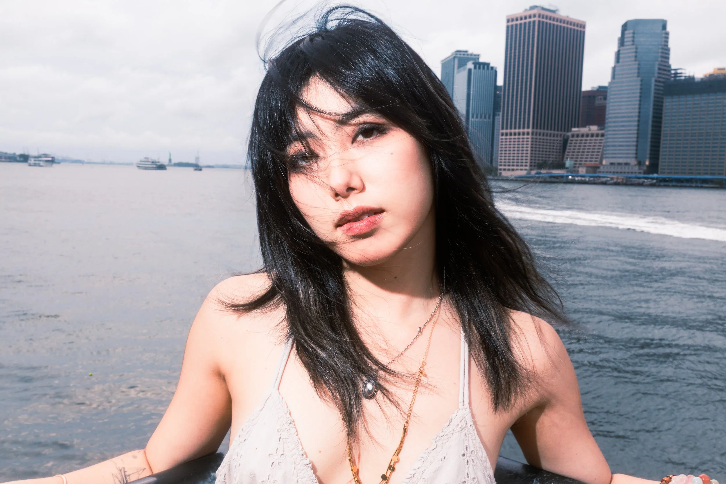 Young woman with black hair and brown eyes on a boat in a city waterfront, with tall buildings and watercraft in the background during overcast weather.