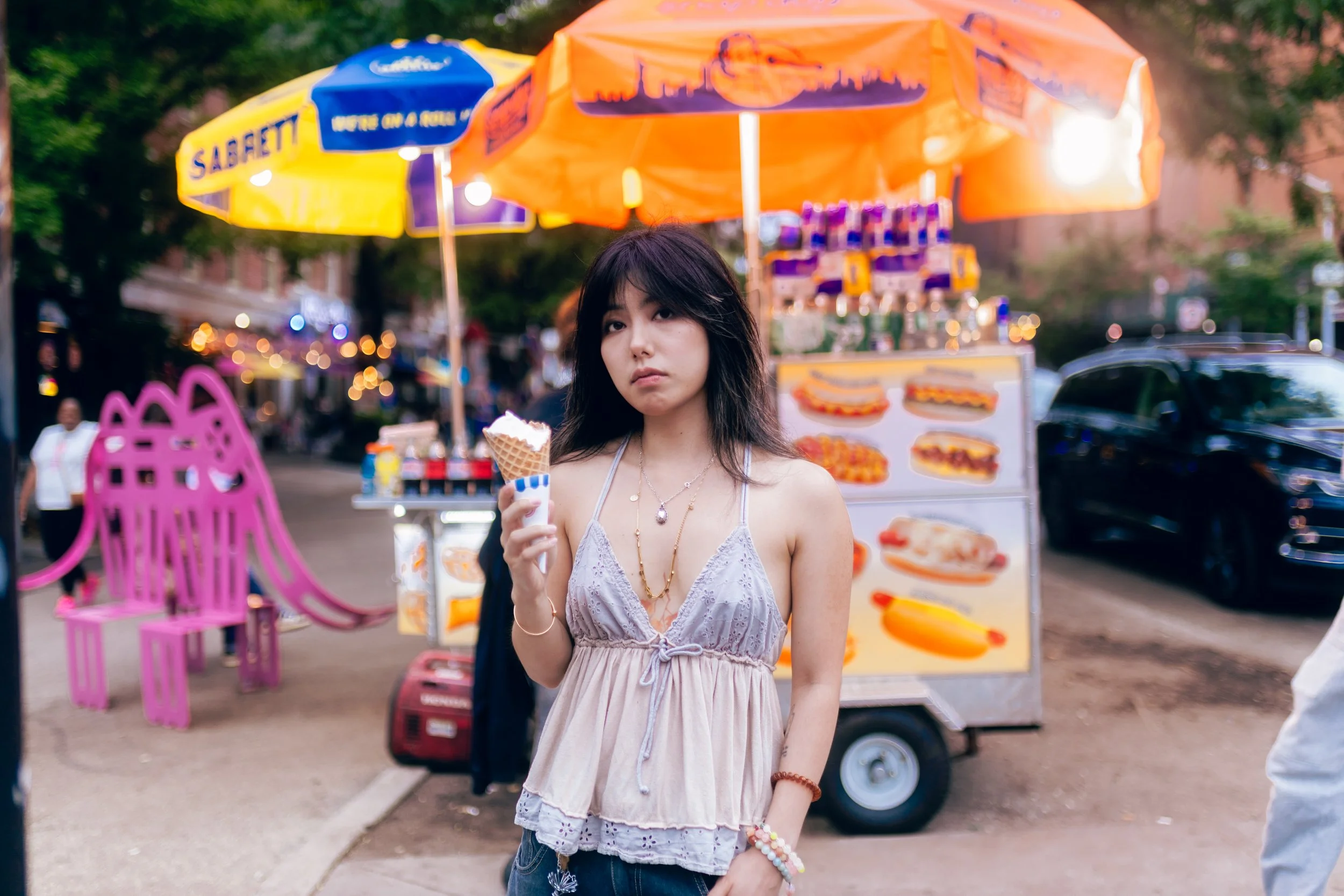 A young woman with dark hair holds an ice cream cone at an outdoor street fair or carnival. She wears a light purple, sleeveless, lace-trimmed top and multiple jewelry, including necklaces and bracelets. Colorful booths and umbrellas, with food poste