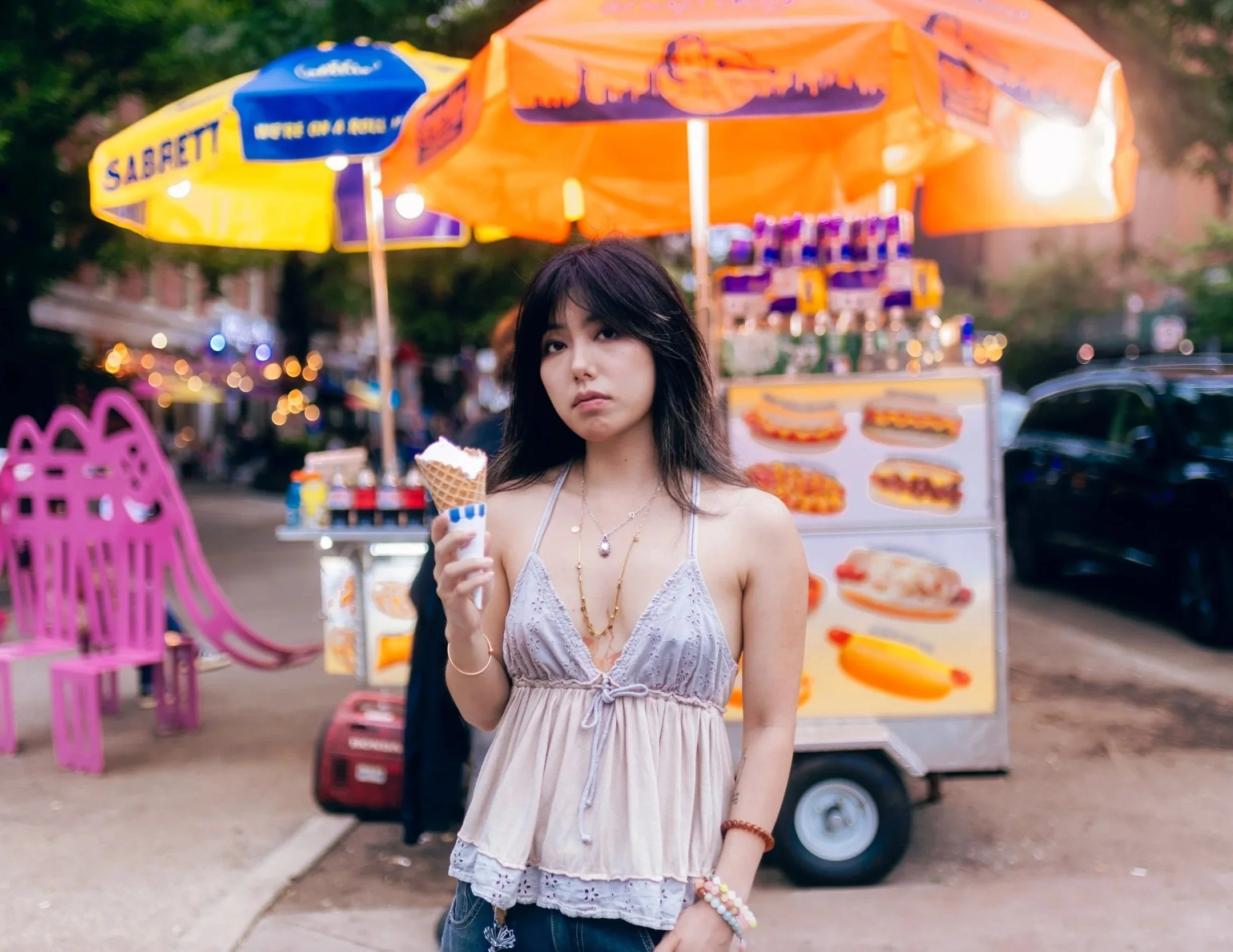 A young woman with dark hair holds an ice cream cone at an outdoor street fair or carnival. She wears a light purple, sleeveless, lace-trimmed top and multiple jewelry, including necklaces and bracelets. Colorful booths and umbrellas, with food poste
