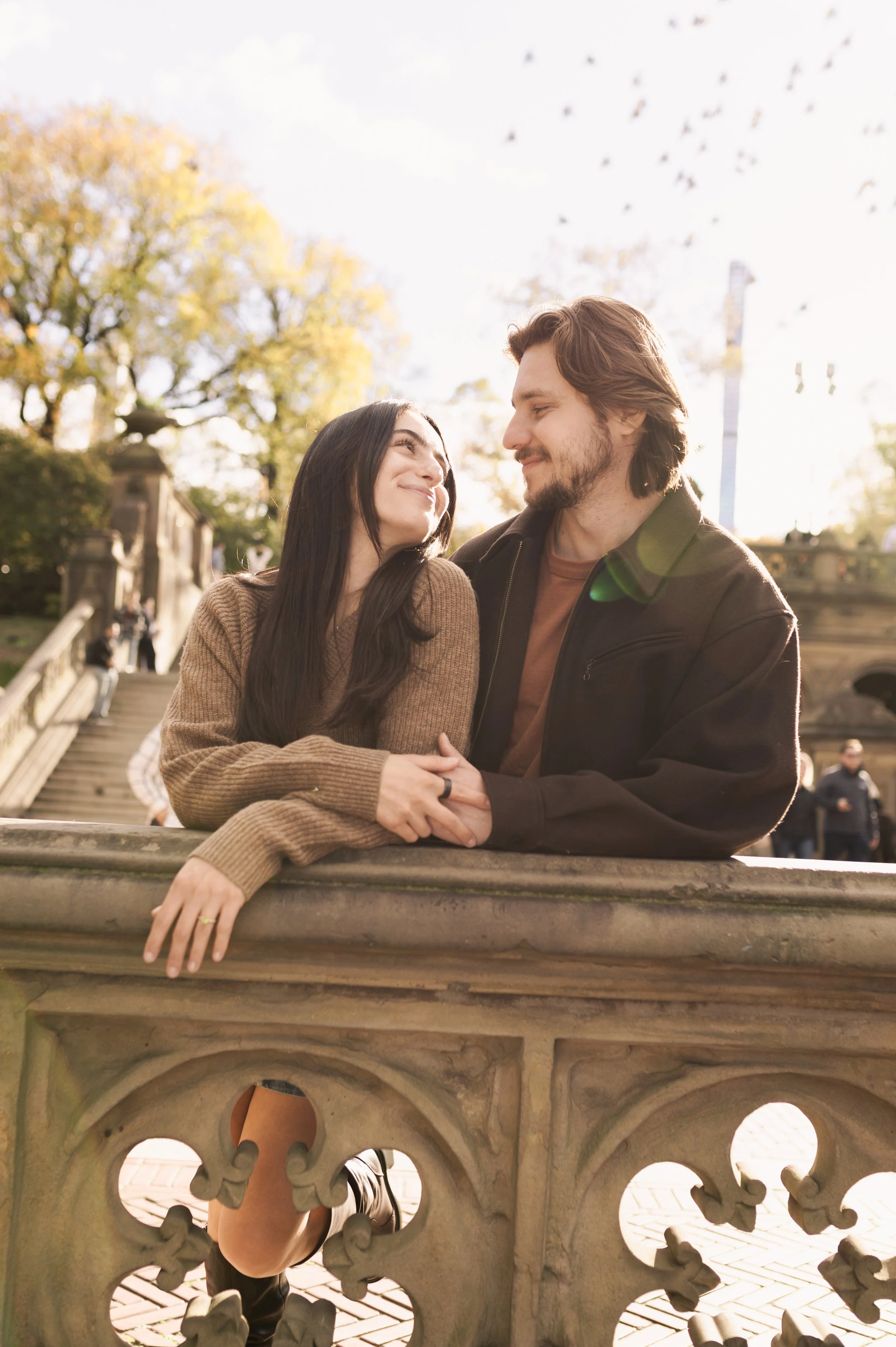 A man and woman sitting on a stone ledge in a park, smiling and looking at each other, with trees and people in the background.