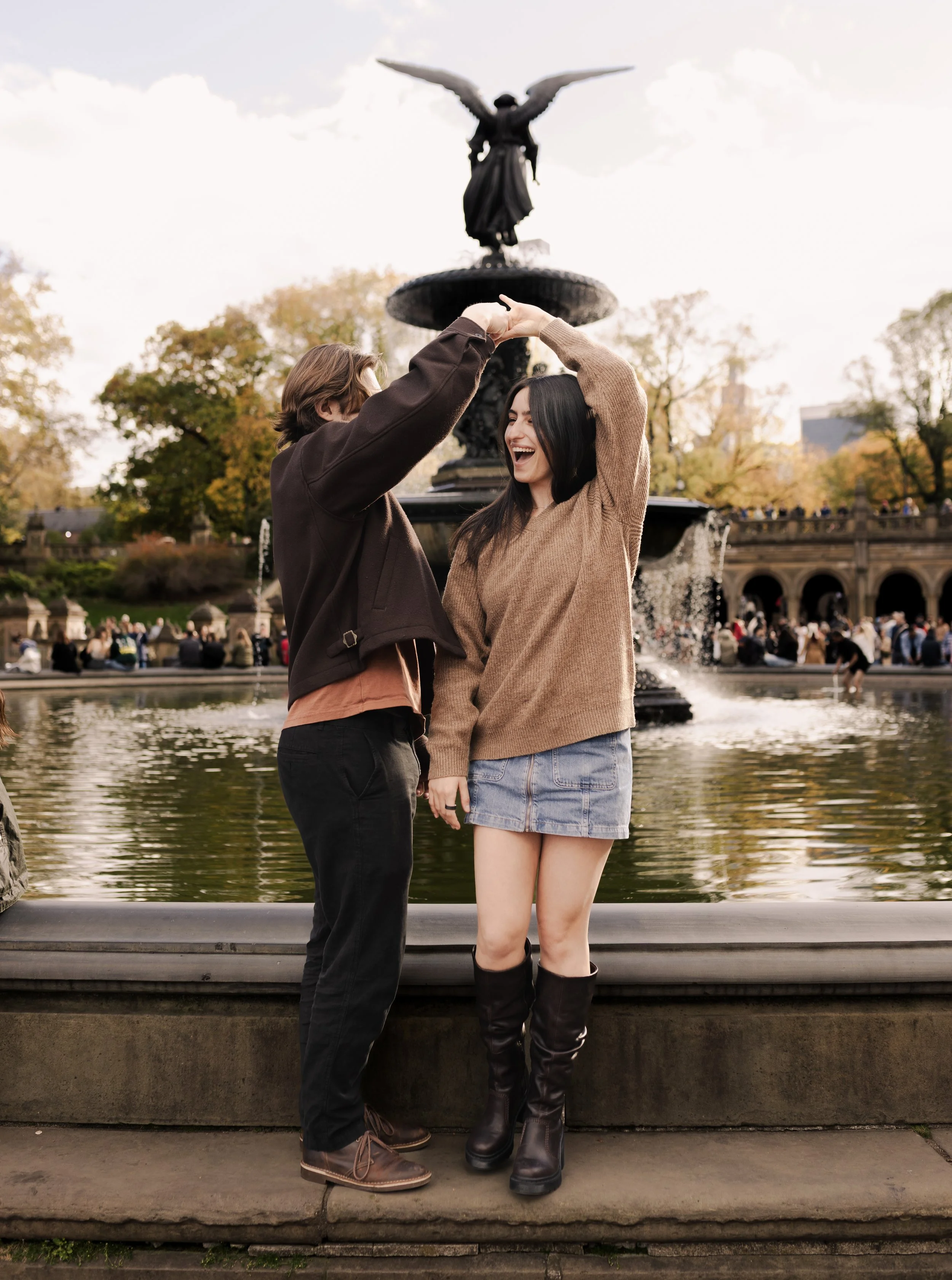 A young couple dancing together outdoors near a fountain in a park with trees and a crowd in the background.