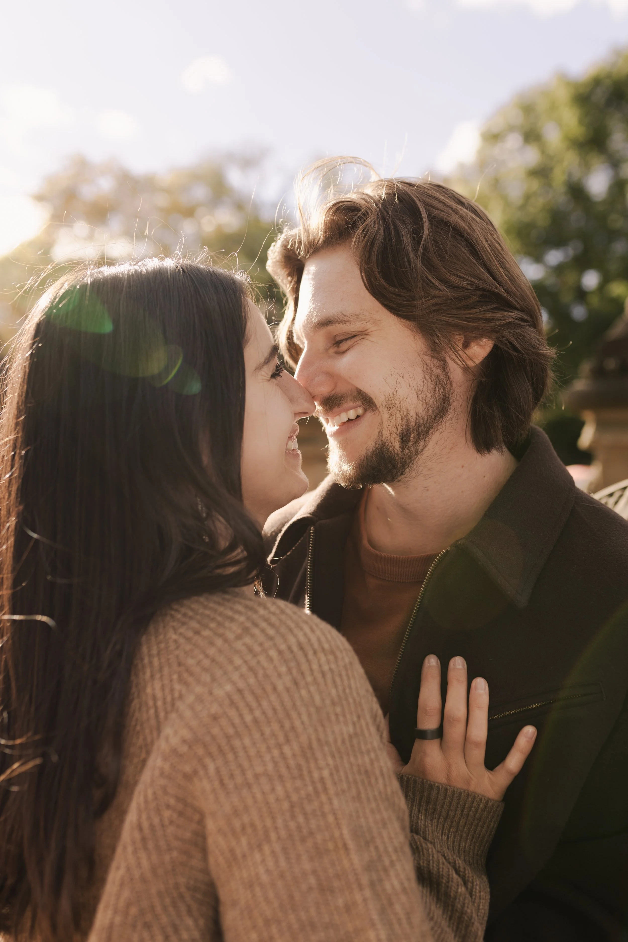 A young couple is smiling and touching noses outdoors in sunlight. The woman has long dark hair and is wearing a brown sweater, and the man has long brown hair, a beard, and is wearing a black jacket.