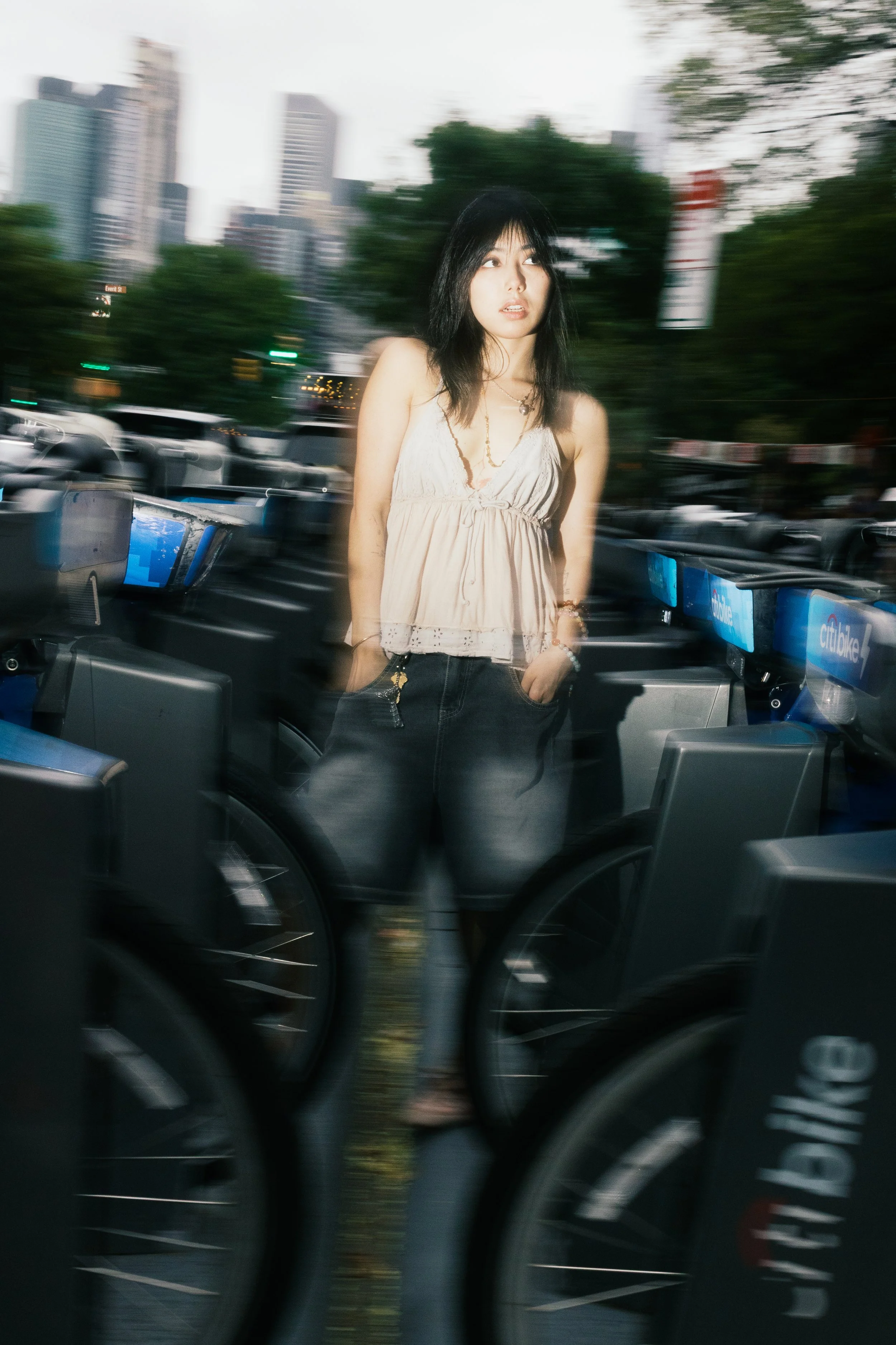 A woman with black hair in a sleeveless beige top and necklace standing at an ATM or kiosk, with a cityscape of tall buildings and trees in the background, and the photo appears to be taken with motion blur.
