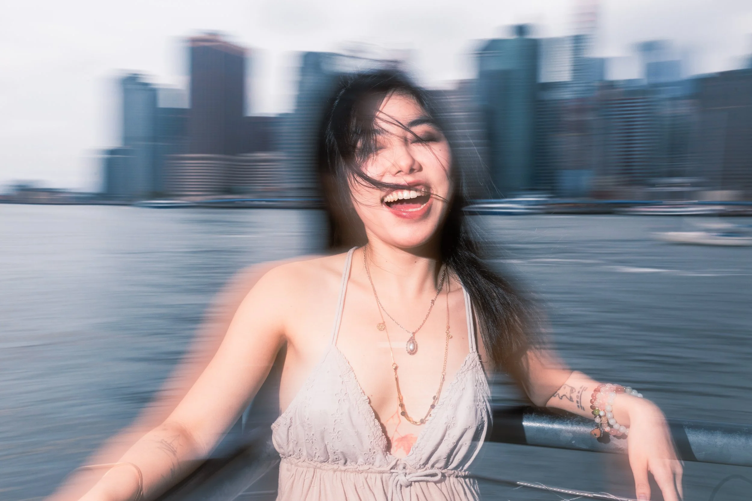 A woman with long dark hair is on a boat, smiling and singing with her eyes closed as wind blows her hair. She is wearing a white lace top, layered necklaces, and bracelets. The background shows a city skyline with tall buildings over water, appearin
