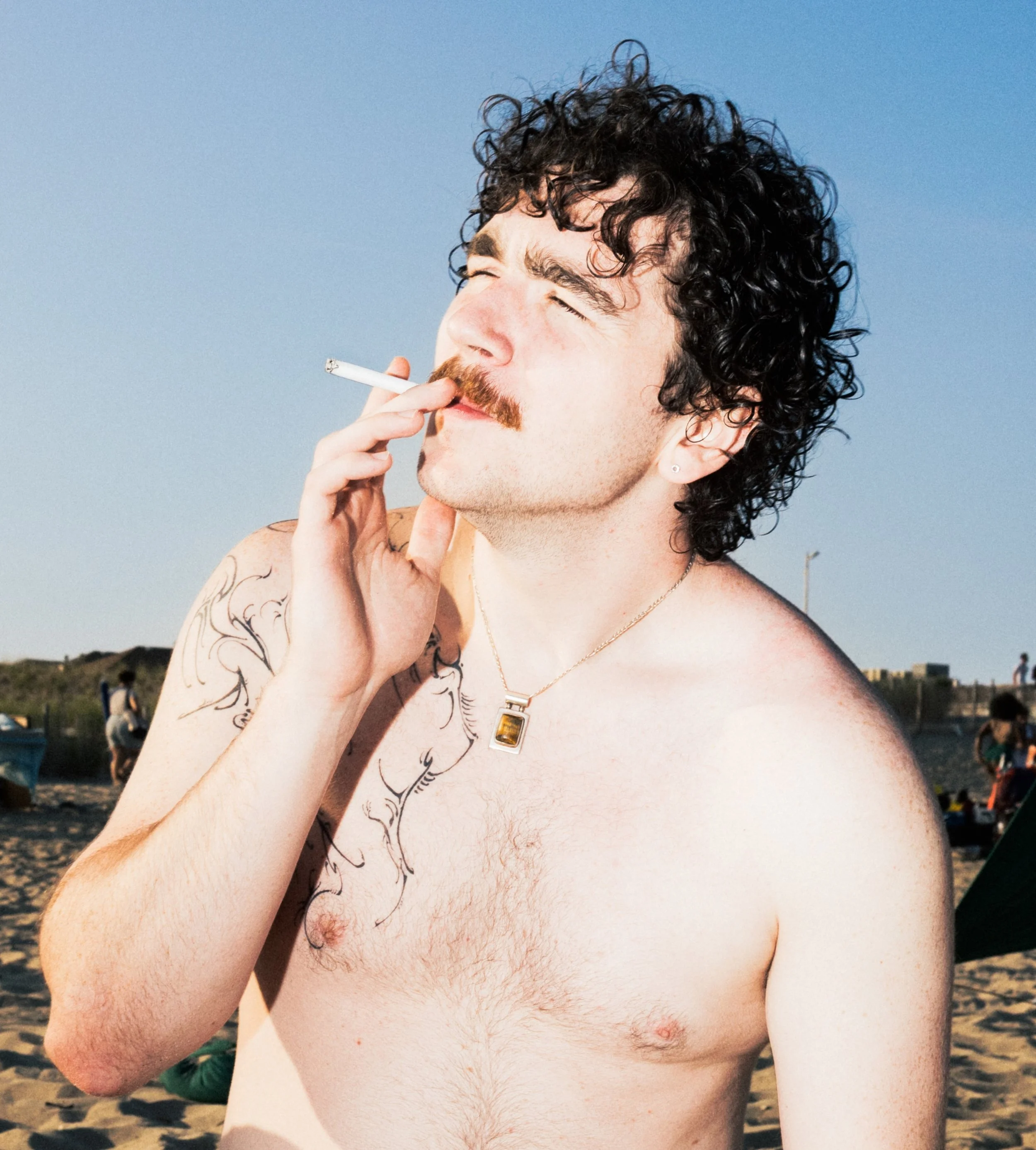 A shirtless man with curly dark hair, a mustache, and tattoos on his arm, holding a cigarette near his mouth, outdoors with a blue sky and a beach background.