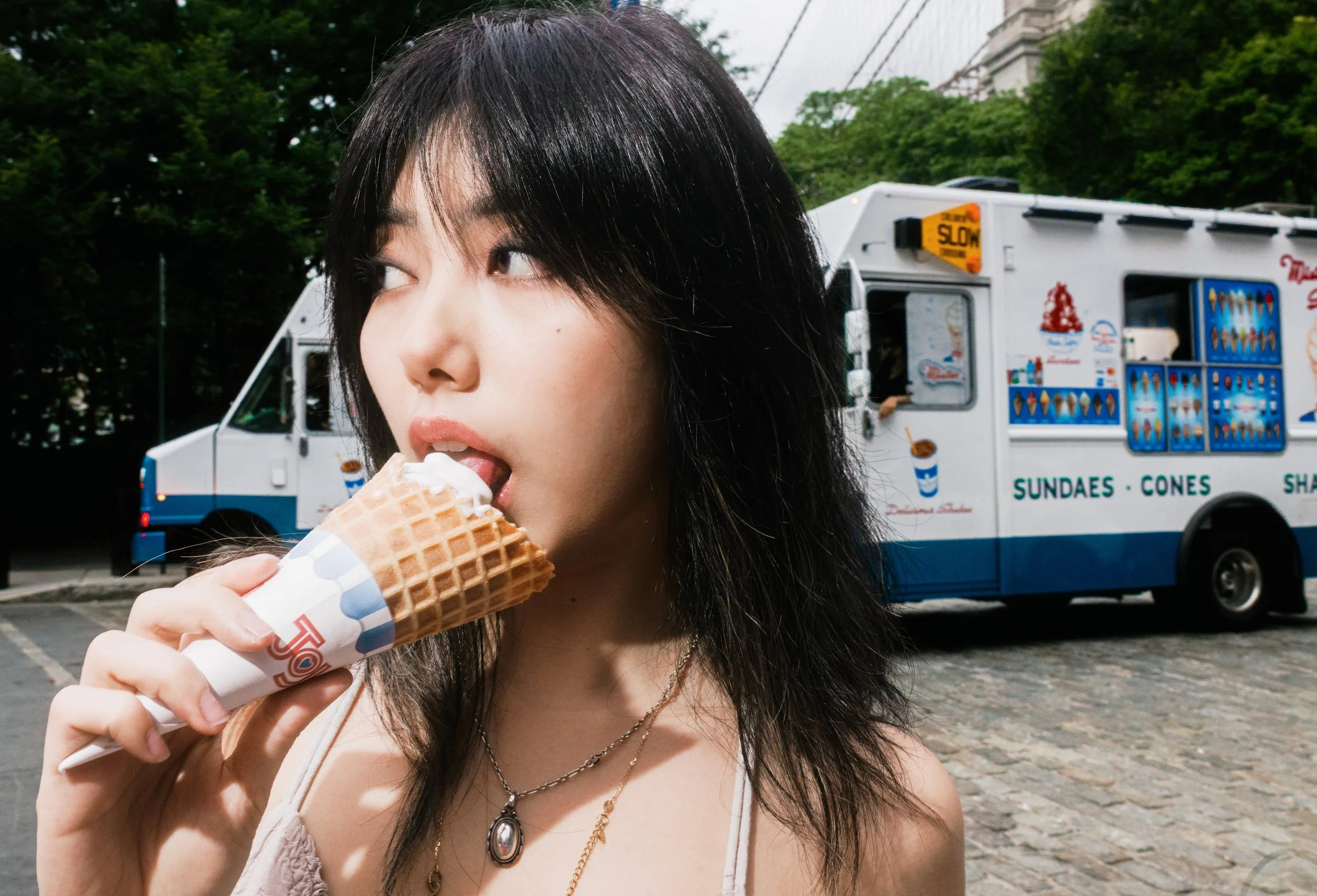 A young woman with dark hair eating an ice cream cone outdoors with a food truck and trees in the background.