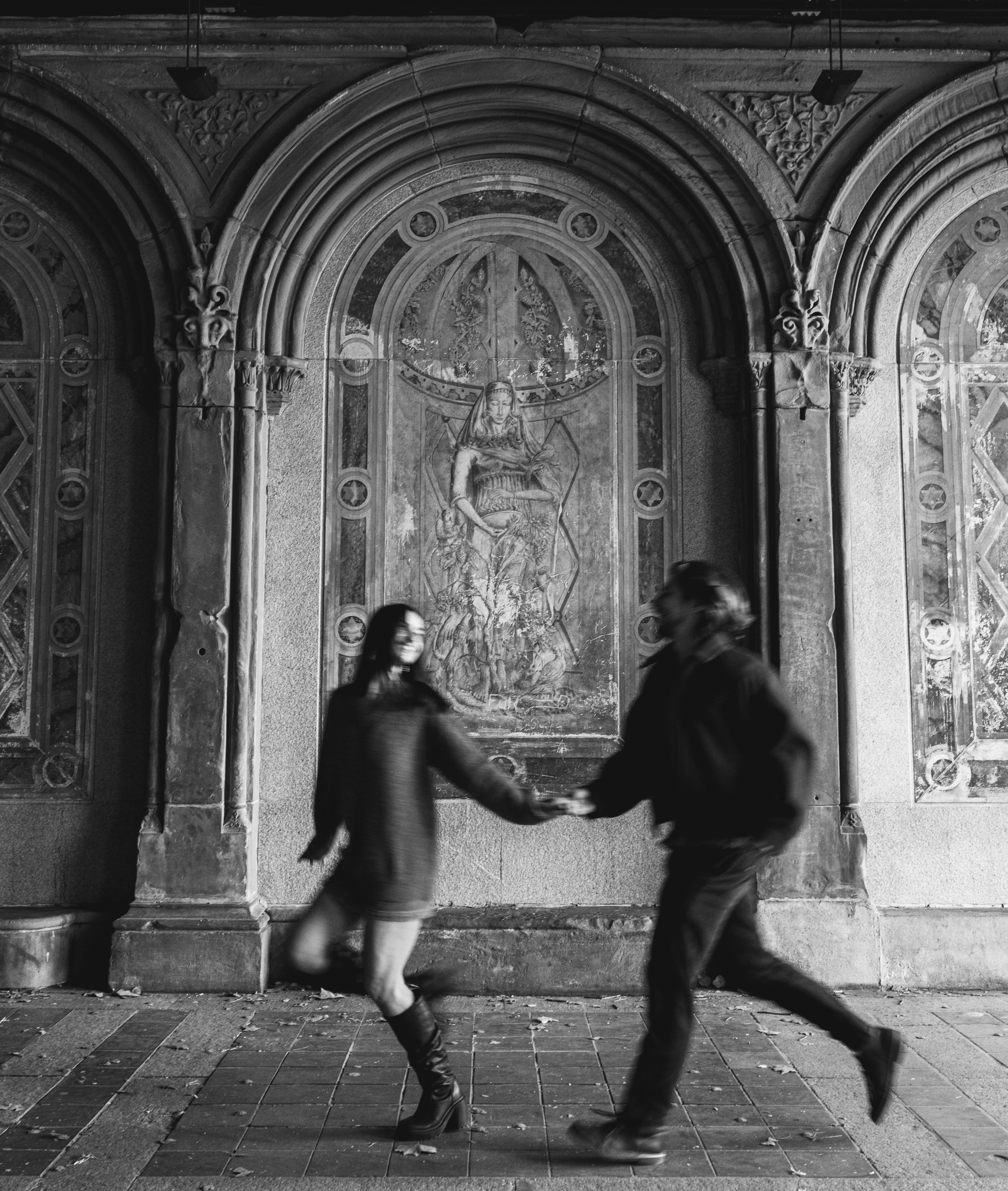 Two people walking past an elaborate stone or concrete archway with detailed carvings and religious imagery, including a statue of a woman holding a sword and a shield, on a city street at night.