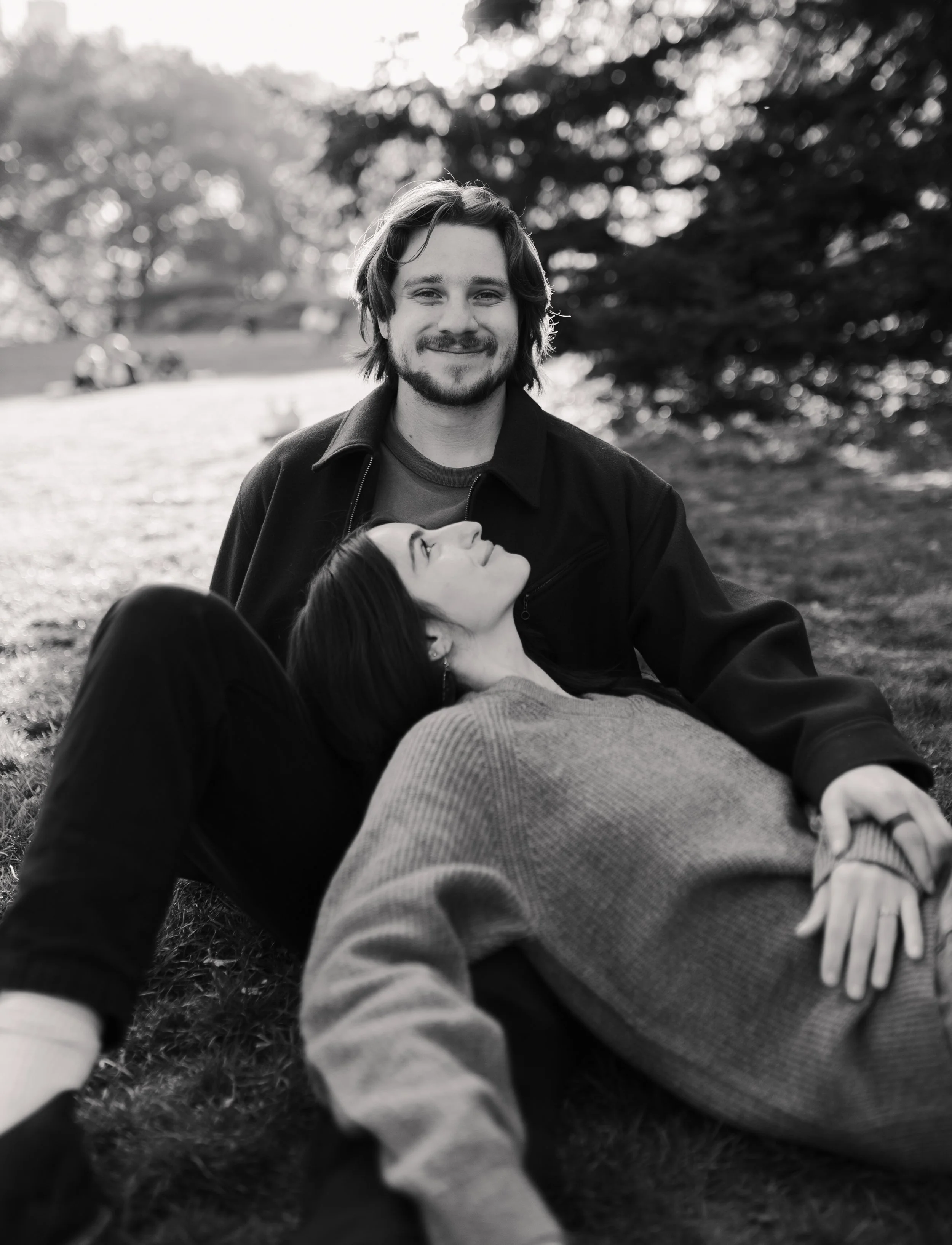 A black and white photo of a young smiling man with shaggy hair and a beard, sitting outdoors on the grass, with a woman laying on his lap looking up at him.