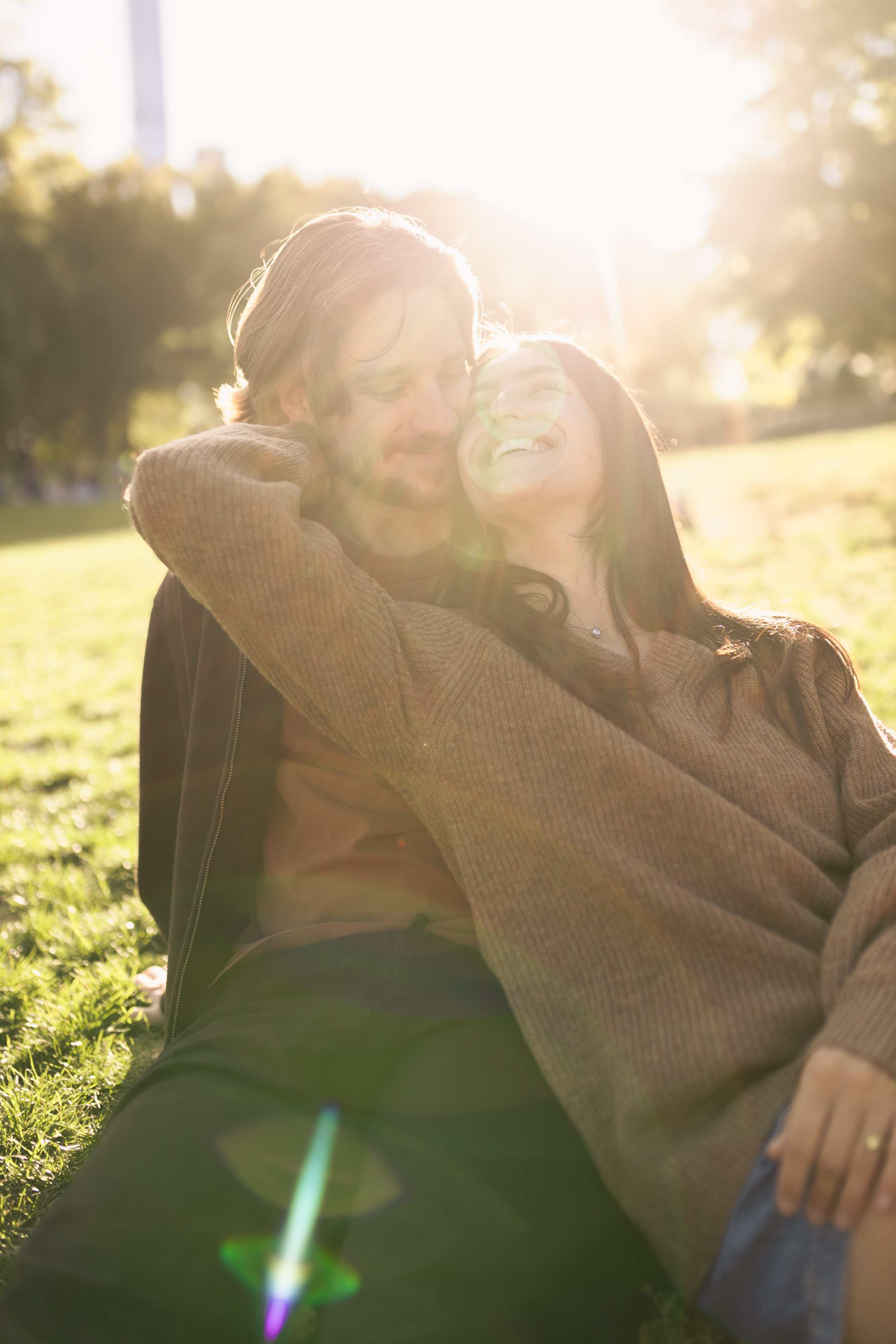 Two people smiling and hugging outdoors with bright sunlight and greenery in the background.
