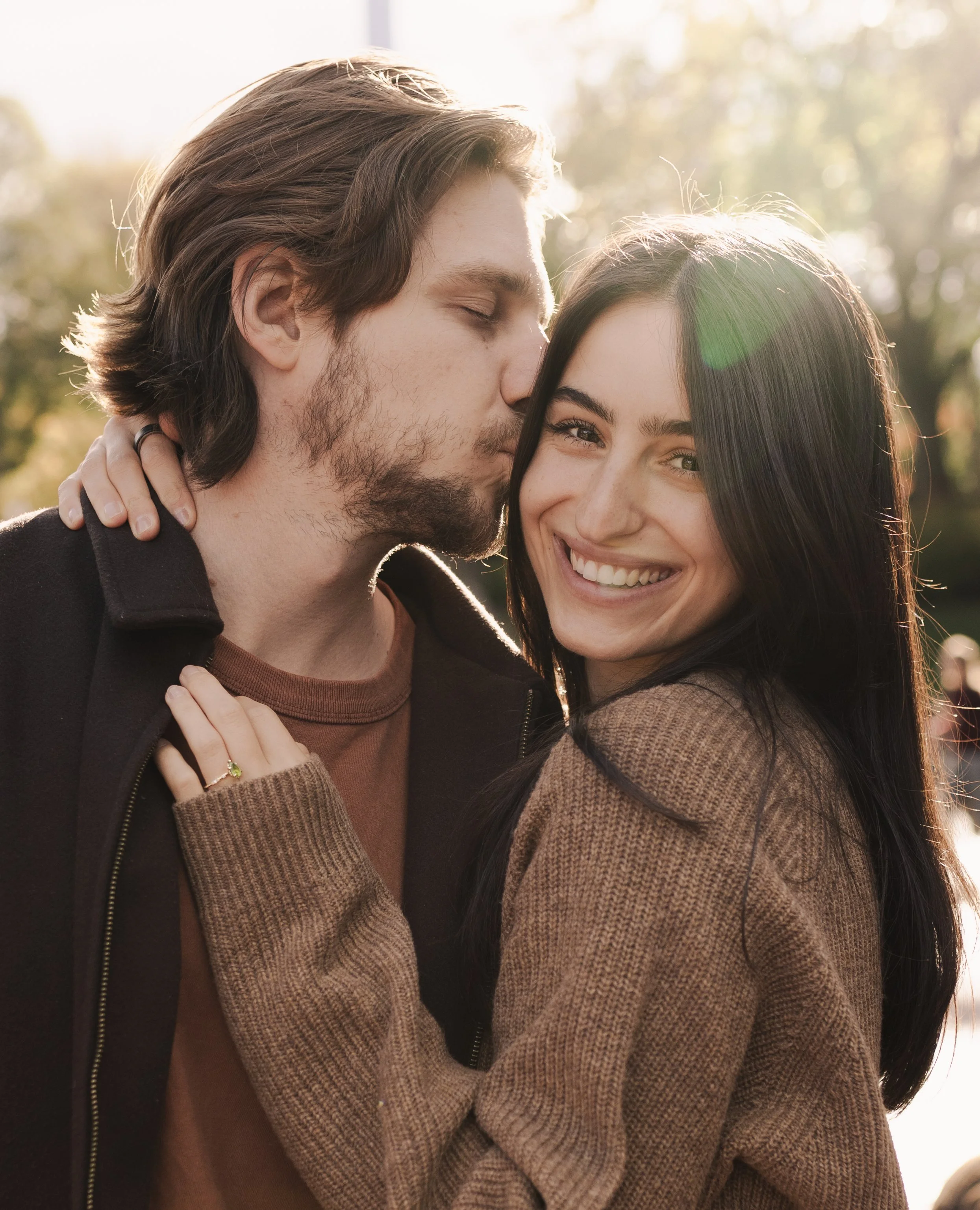 A man and woman enjoying a close, affectionate moment outdoors. The man is kissing the woman on the forehead, and they are smiling happily. The background features sunlight filtering through trees.