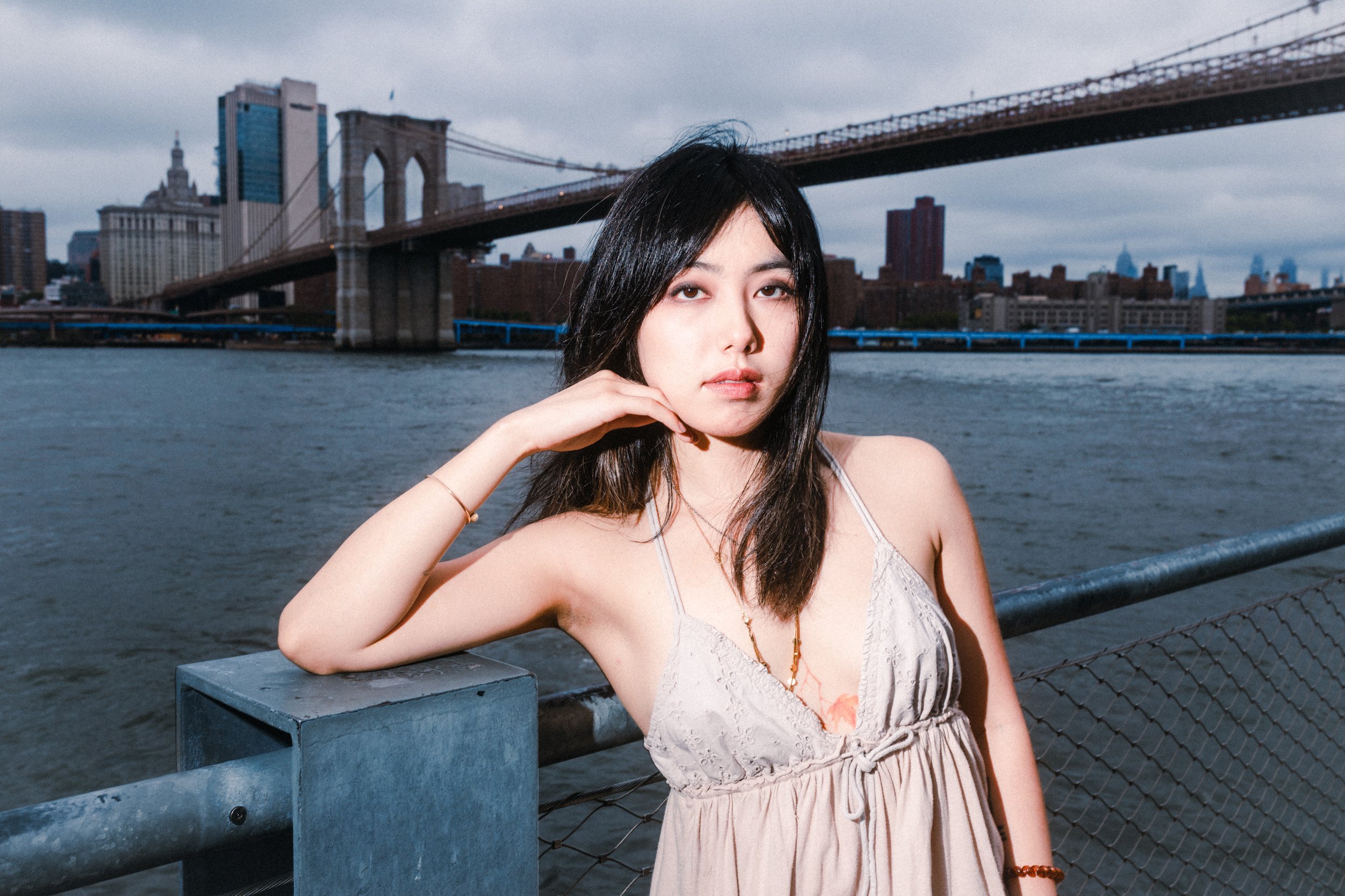 A young woman with dark hair, posing against the backdrop of the Brooklyn Bridge and NYC skyline along the river, wearing a light-colored dress and jewelry.