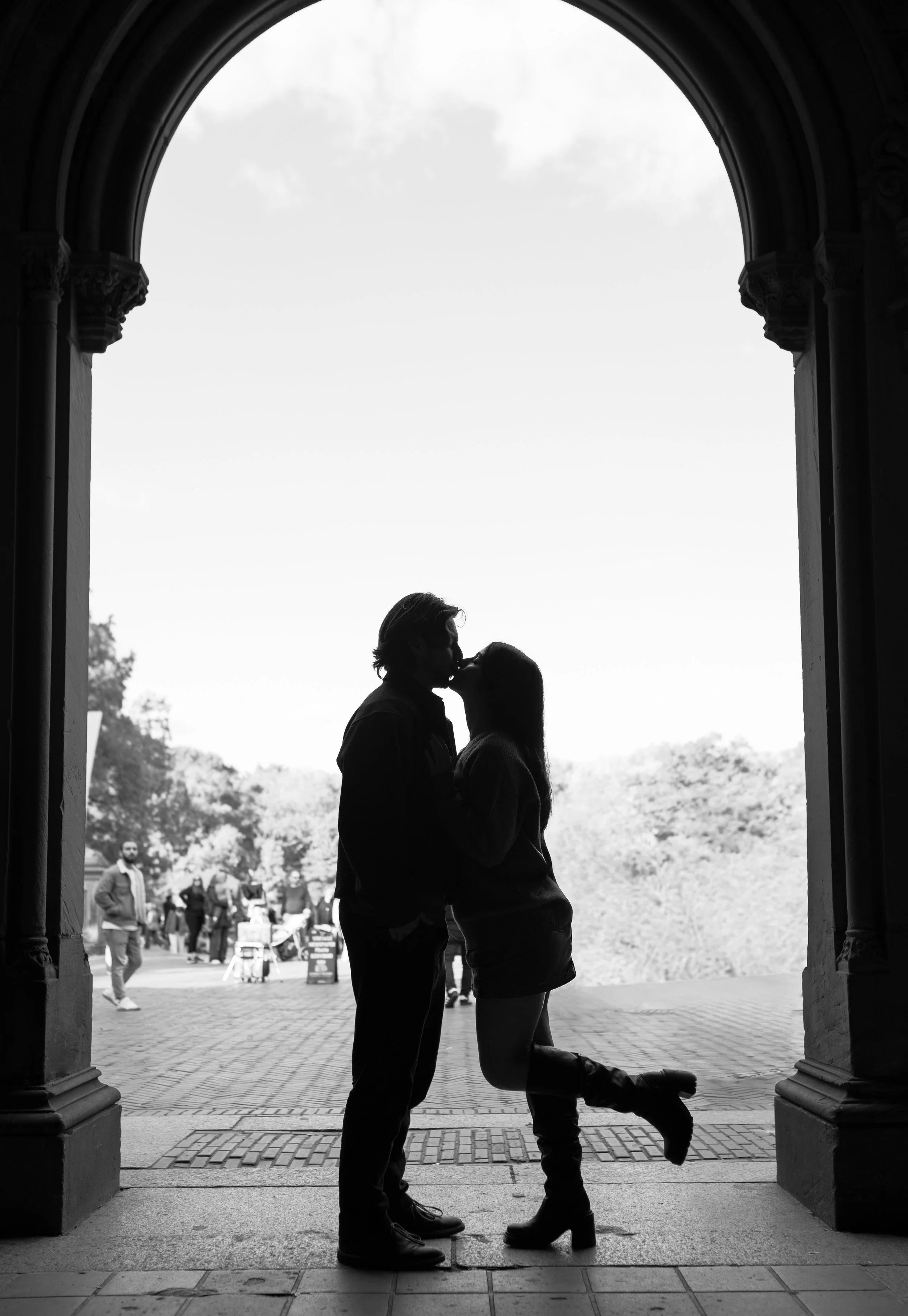 Silhouetted couple kissing under an archway, with other people and trees outside in the background.