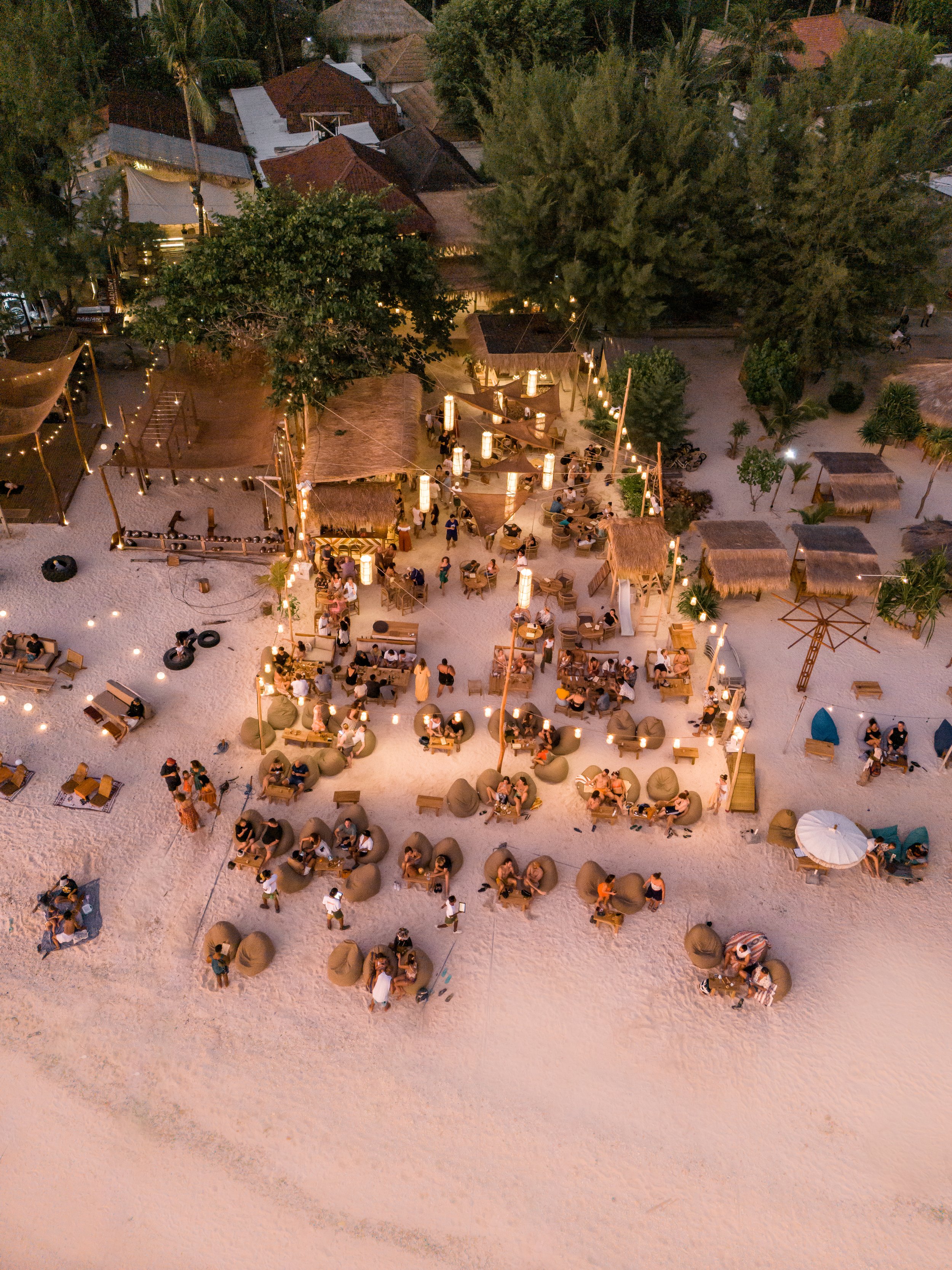 Aerial view of a beachside venue at dusk, with groups of people sitting on bean bags and chairs, illuminated by string lights, with tiki huts and trees around.