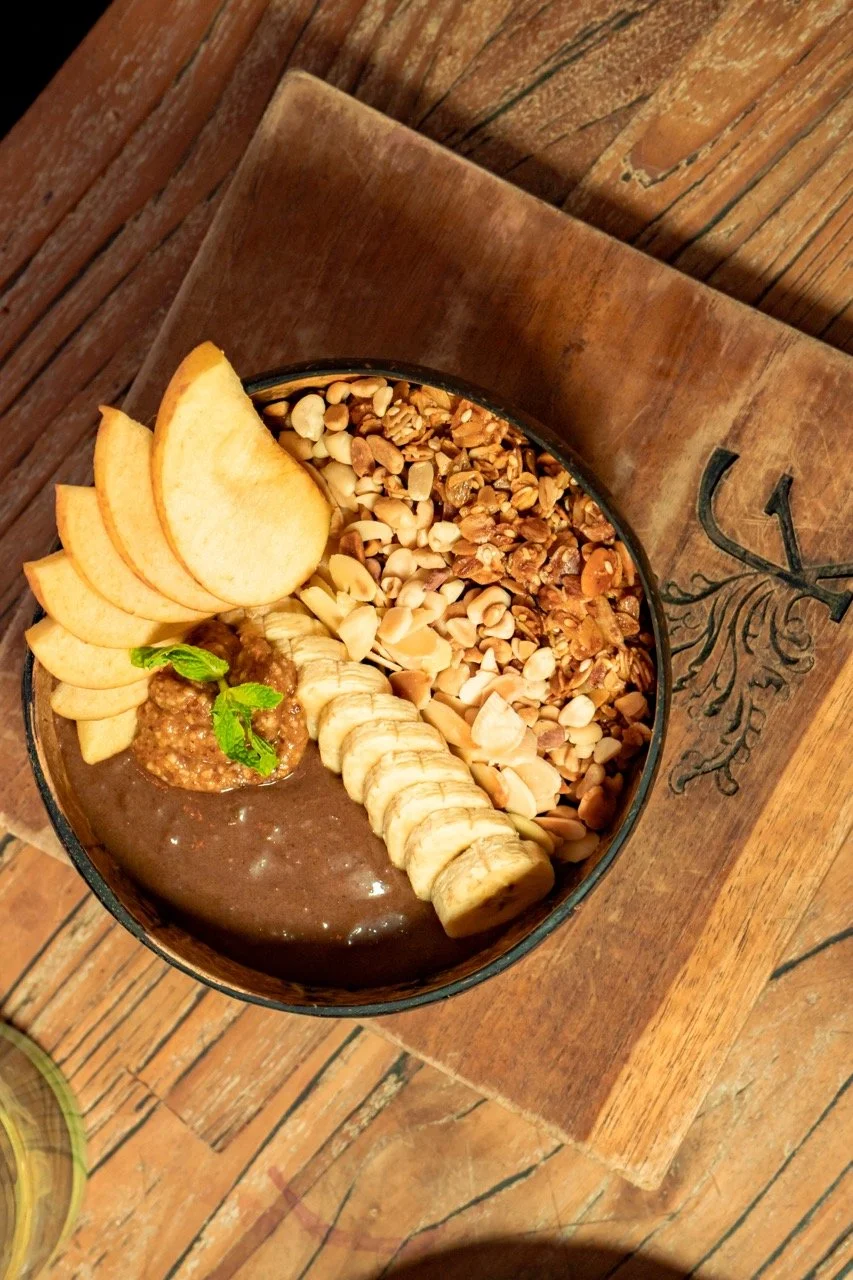 A bowl of chocolate chia pudding topped with sliced banana, sliced apple, granola, chopped nuts, and a sprig of mint, placed on a wooden table.
