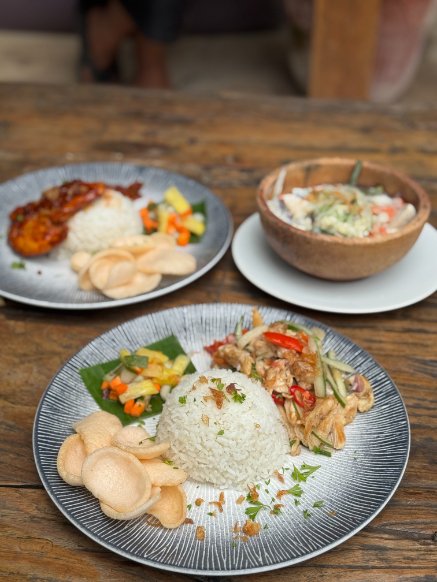 Three plates of food on a wooden table, featuring rice, vegetables, chicken, and a bowl of salad.