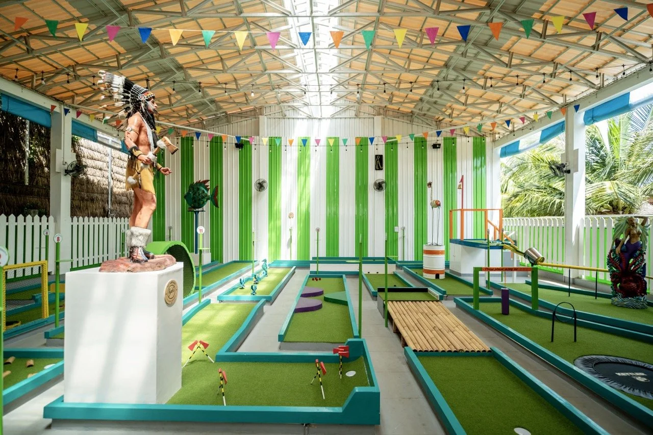 Indoor miniature golf course with various themed holes, colorful bunting hanging from the ceiling, and statues of a Native American chief and a woman, surrounded by tropical plants outside.