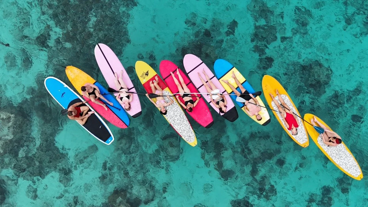 Aerial view of seven people paddleboarding on clear turquoise water, with some wearing swimsuits and others in cover-ups, on colorful paddleboards.
