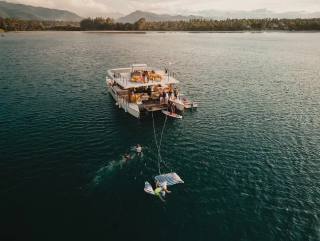 A large white catamaran on the Gili waters with people on the deck, towing a floating mattress with a person lying on it, while a boat pulls the mattress.