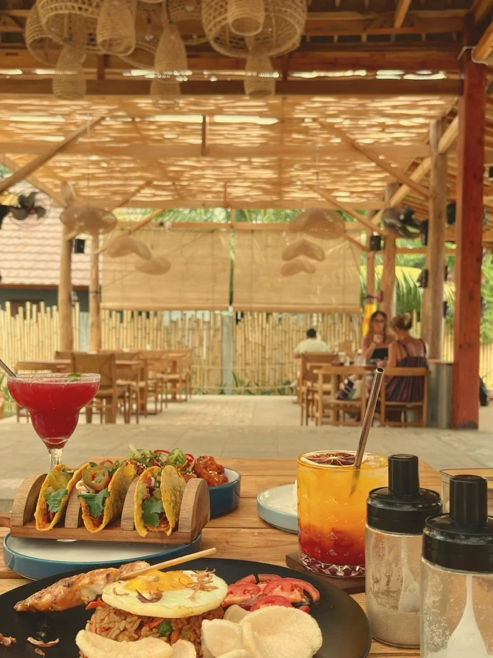 A table with Mexican tacos, grilled chicken, and rice, next to drinks including a red cocktail, an orange-yellow beverage, and a pinkish cocktail, in an open-air restaurant with bamboo decor and two women dining in the background.