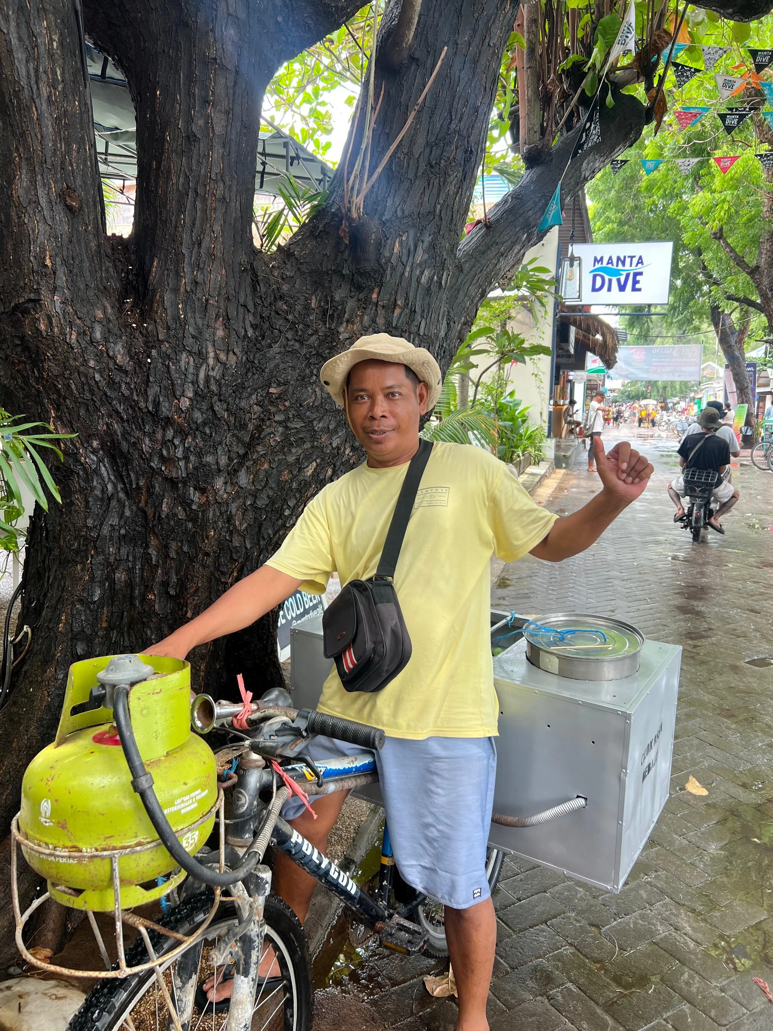 A man in a yellow t-shirt and blue shorts uses a bicycle equipped for selling ice or cold drinks on a rainy street. He is standing near a large tree and pointing at a sign that says 'Manta Dive.'
