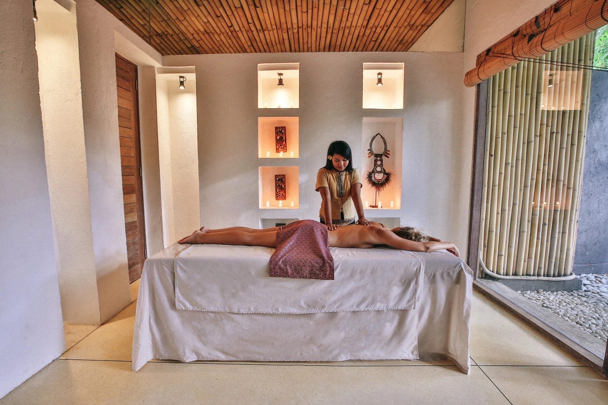 A woman receiving a massage on a massage table in a spa room with soft lighting, candles, and decorative wall niches.