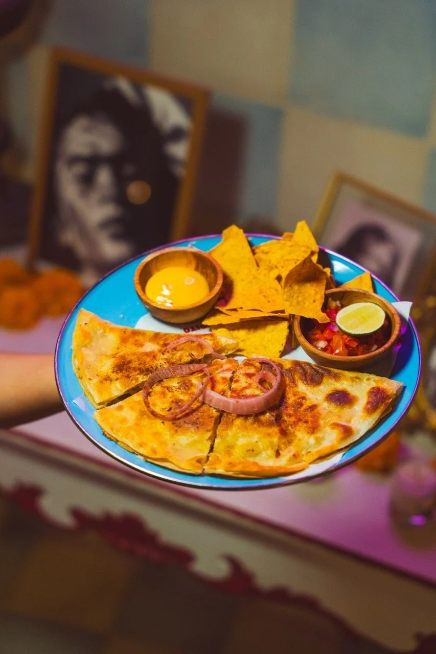Plate of cheese pizza with onion slices, tortilla chips, and two small bowls of salsa and mustard, with a lime wedge in the salsa, against a background of framed black-and-white photographs.