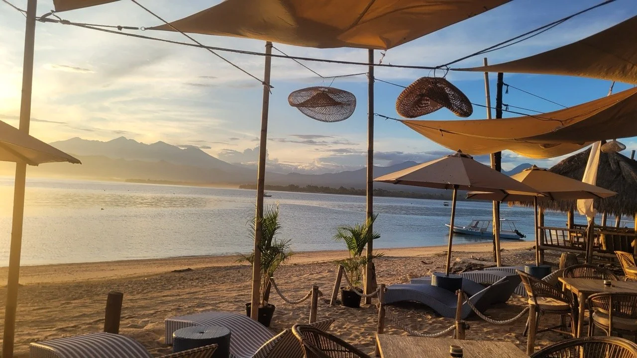 Beachside outdoor seating area with umbrellas and lounge chairs at sunset, overlooking a calm river with mountains in the background.