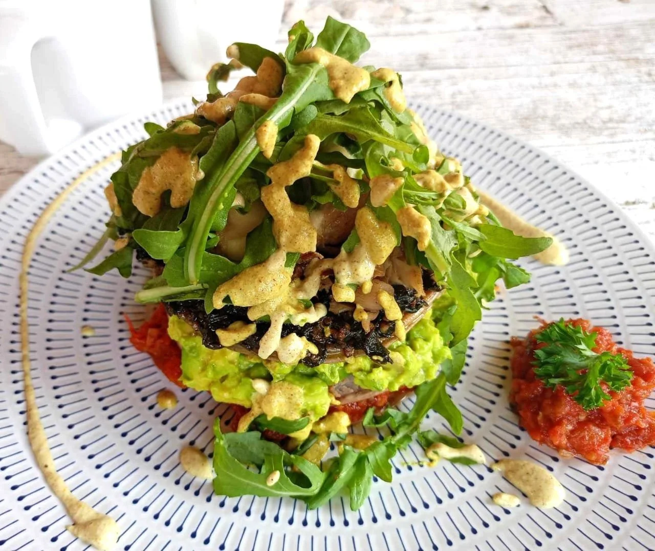 A plated taco with guacamole, sliced vegetables, topped with leafy greens, crispy cheese, and garnished with cilantro, served with side of salsa and mustard sauce on a white plate.