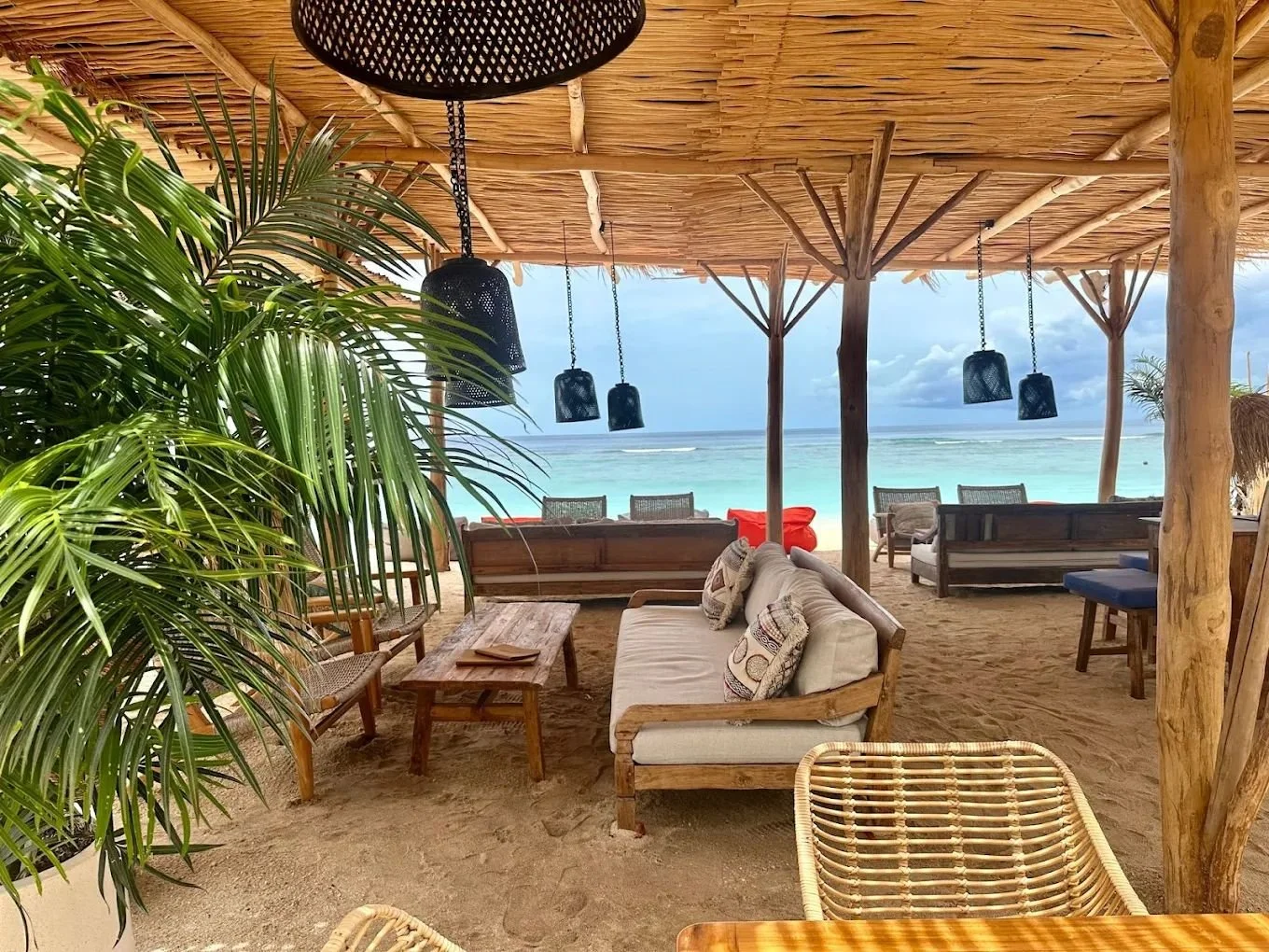 Beachside lounge area with wooden furniture, cushions, hanging lamps, and a view of the ocean and sky.