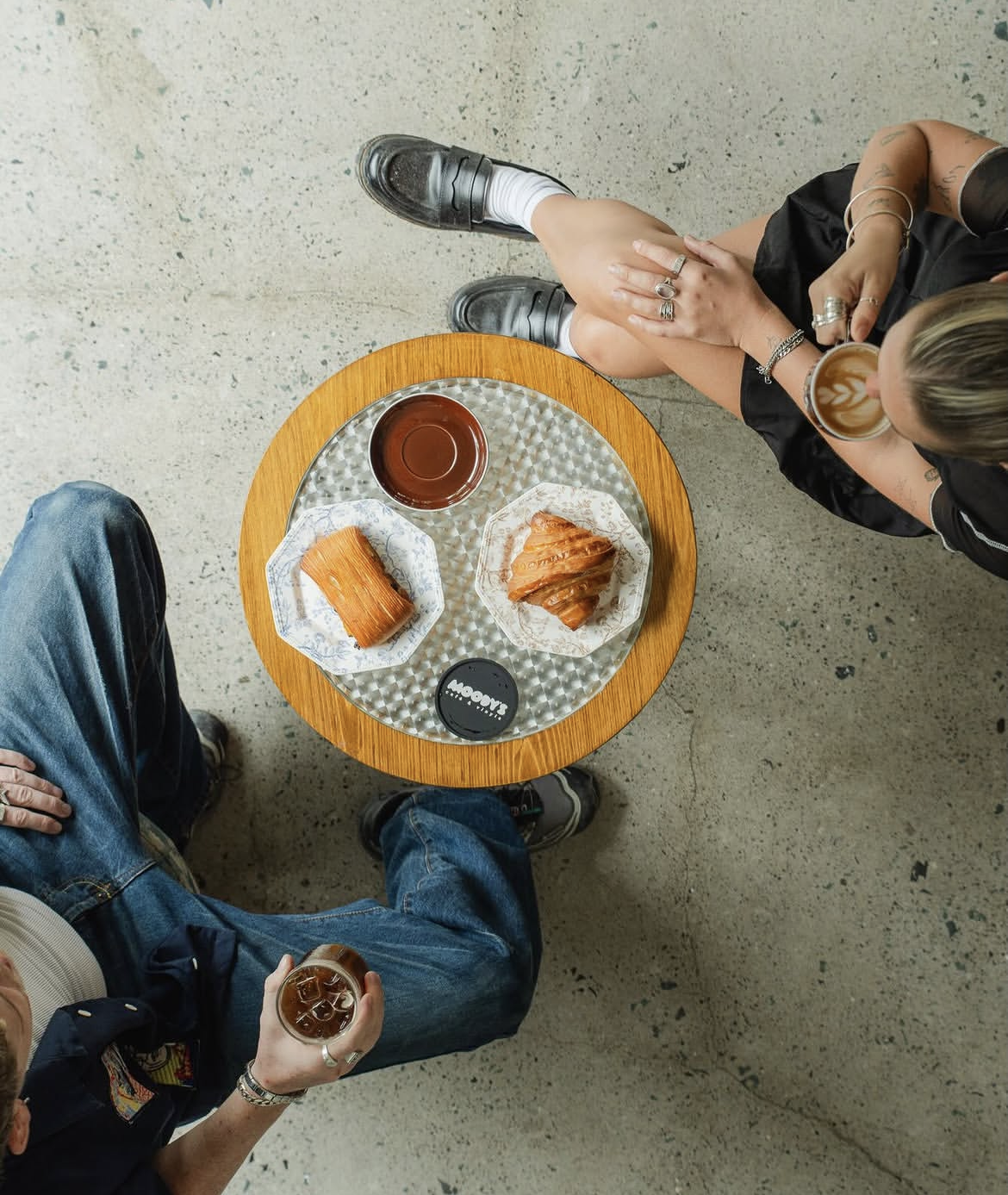 Top-down view of a small round wooden table with two croissants, a glass of tea, and a cup of coffee, with two people sitting around it, one holding a glass of iced coffee.