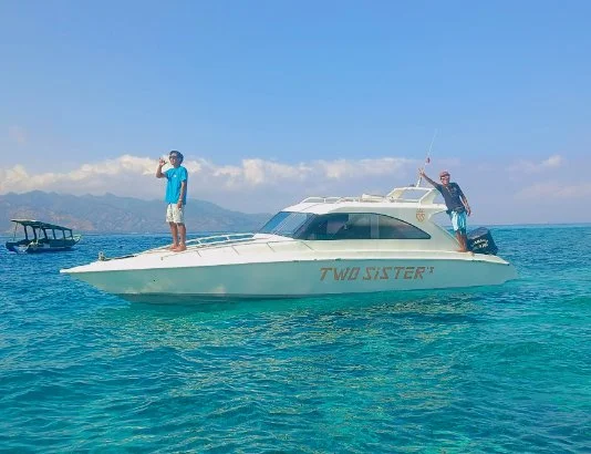 Two men on a white boat named 'TWO SISTER' in clear blue ocean waters, with mountains and clouds in the background.