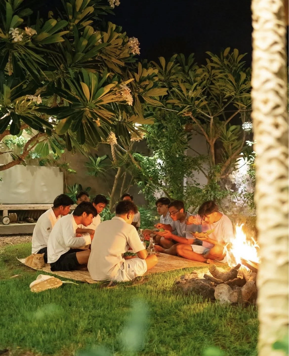 Group of young people sitting on a mat around a campfire at night, eating outdoors under trees