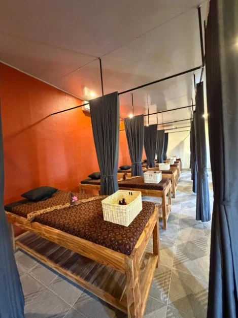 A row of four beds with dark brown mattresses and pillows, separated by privacy curtains in a spa or relaxation room.