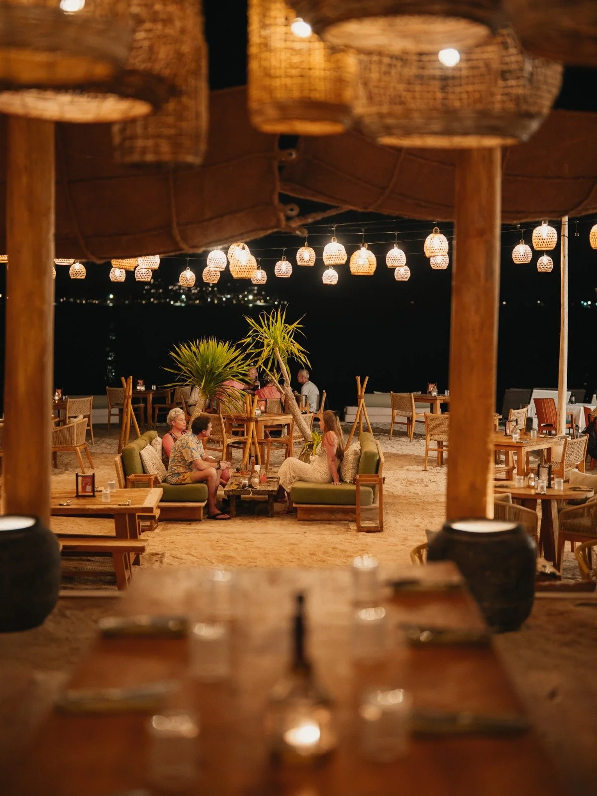 Outdoor restaurant at night with hanging wicker lamps, wooden tables, chairs, and a few groups of people dining under a large canopy.