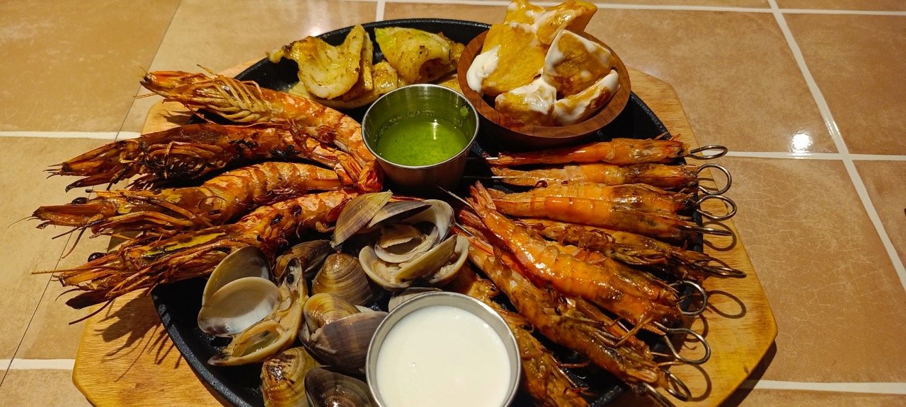 Seafood platter with grilled shrimp, clams, and several sauces, served on a wooden tray.