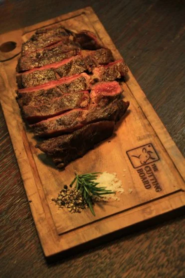 Sliced cooked steak with herbs, garlic, and pepper on a wooden cutting board.