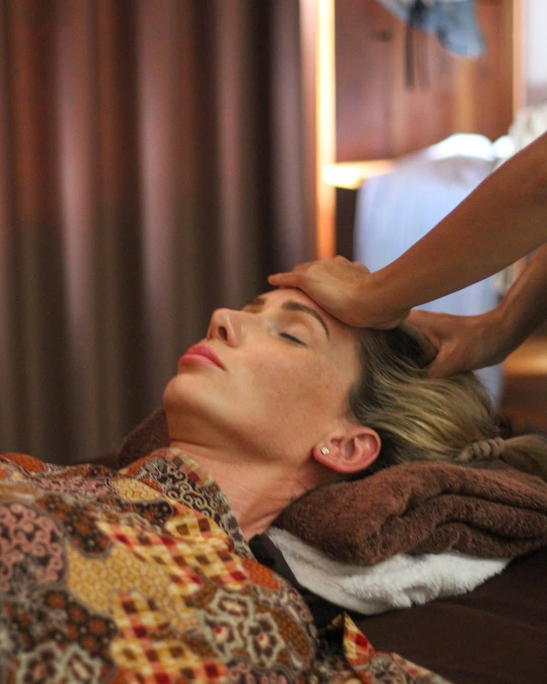 A woman lying on her back with eyes closed, receiving a head massage in a cozy, warmly lit room with wooden accents.
