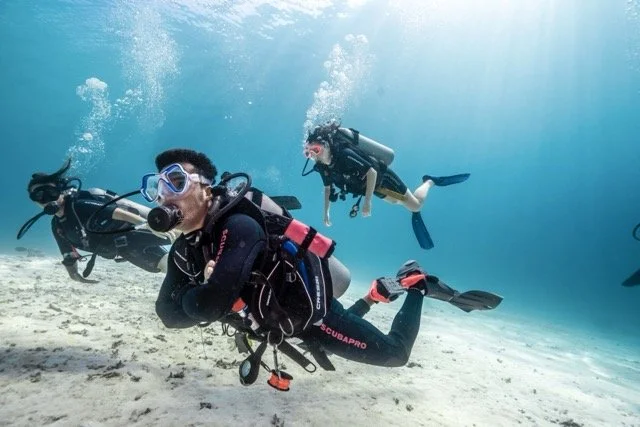 Two scuba divers exploring underwater, one in the foreground and another in the background, surrounded by clear blue water and sandy seabed.