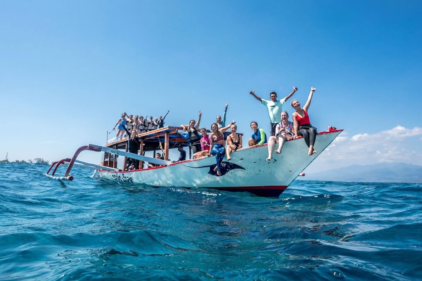 A group of people on a boat in the ocean, with some sitting on the edge and others standing, waving and smiling under a clear blue sky.